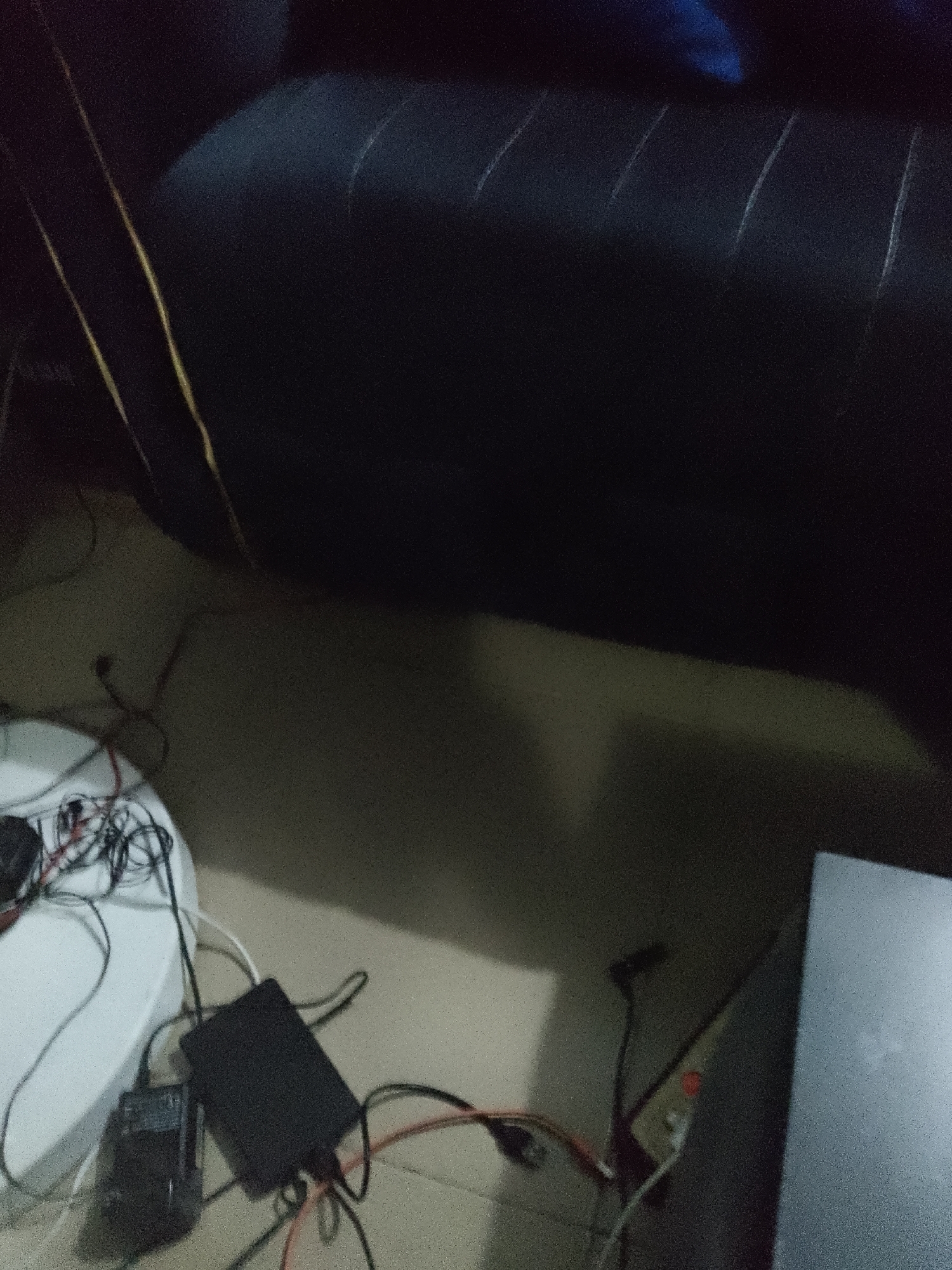 The image shows a dark room with a black couch in the background. In the foreground, there is a white circular object with wires and electronic components on it. There is also a black rectangular electronic device on the floor, connected to various wires. To the right, a portion of a grey electronic device is visible. The lighting is dim, and the scene appears to be indoors, likely in a residential setting in Unknown, Unknown. There are no people or discernible actions taking place.