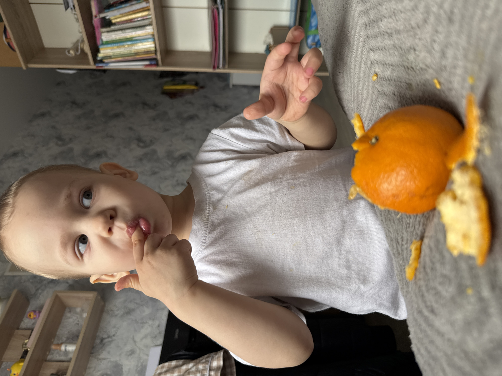 A young child with blond hair is holding a finger to their mouth while looking at the camera. A partially peeled orange is on a gray surface near the child. The location appears to be indoors, near shelving and patterned wallpaper. The city is Rusanovo, Russia.