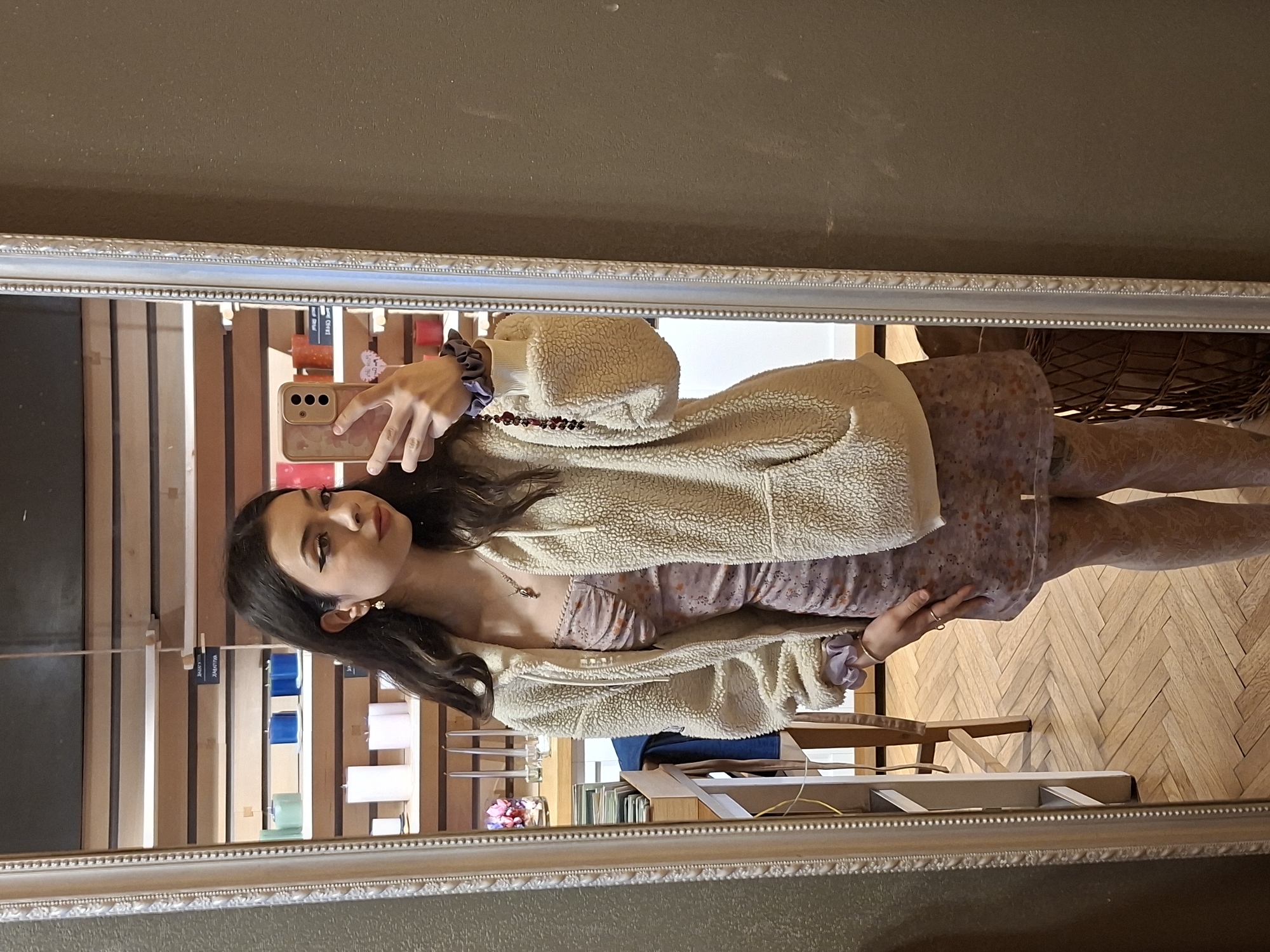 The image shows a young woman taking a selfie in a mirror. She is wearing a floral dress, a cream-colored sherpa jacket, and tights. She has long, dark hair and is holding a smartphone. The reflection in the mirror shows a display of candles and a view of a room with wooden floors and a chair. The location is possibly in Sofia, Bulgaria, as the context suggests.