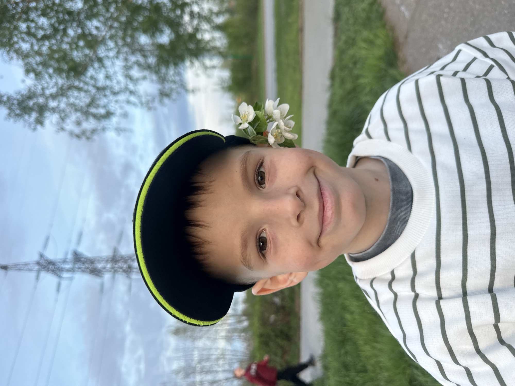 A young boy wearing a black baseball cap with a neon green brim and a white and gray striped shirt is smiling at the camera. There are white flowers tucked into the side of his cap. The background shows a sidewalk, grass, and trees. In the distance are power lines and a person wearing a red jacket. The photo was taken outside in what appears to be the city of Chaikovsky, Russia.