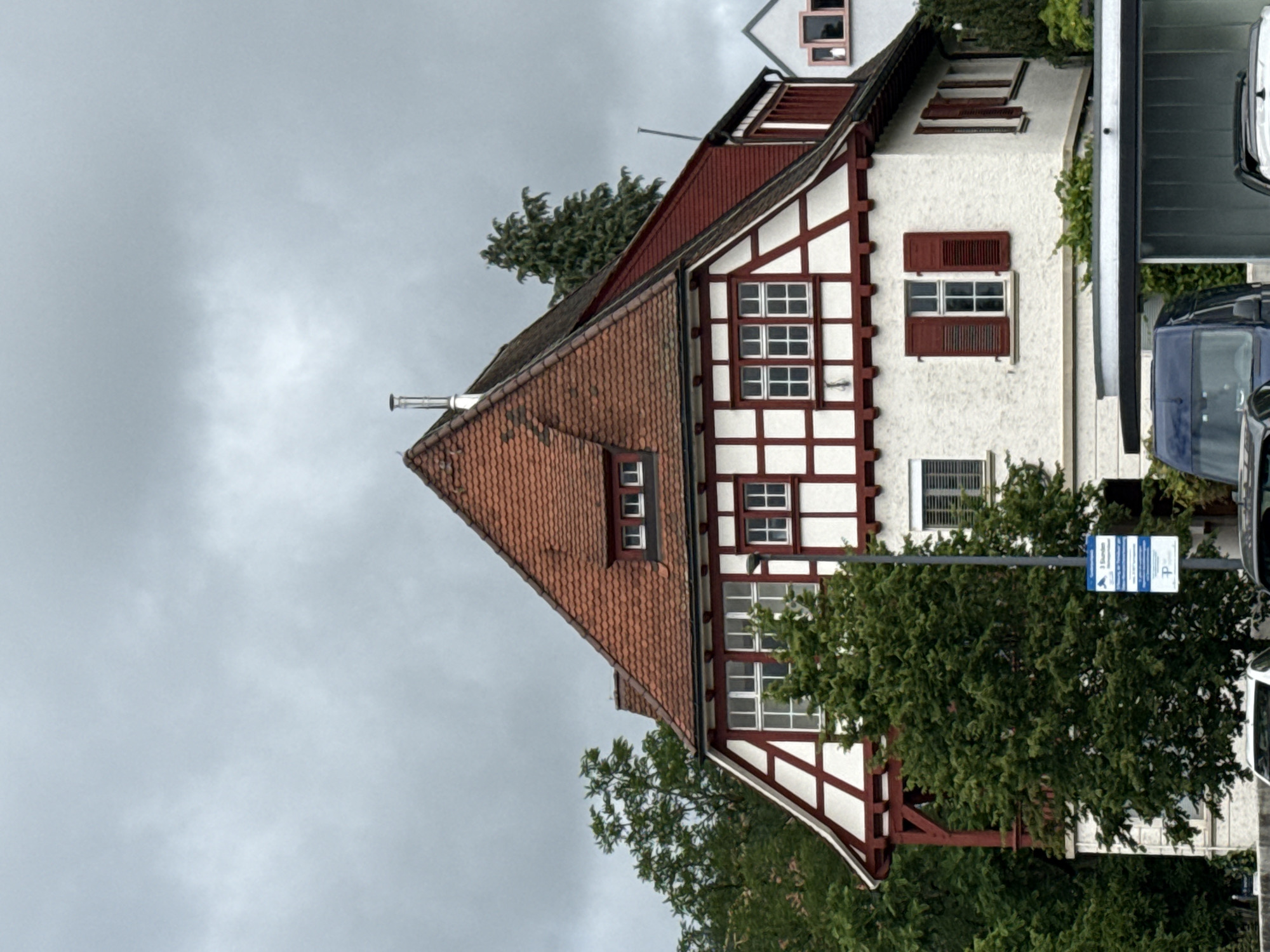 An image taken in Waldshut, Germany, captures a traditional German house with a half-timbered facade under an overcast sky. The house, situated on the left side of the frame, features white stucco walls with prominent dark red wooden beams forming a half-timbered pattern. It has a steeply pitched roof covered in reddish-brown clay tiles, with several small dormer windows protruding from the roofline. A metallic chimney is visible near the peak of the roof.
On the upper floor of the house, there are windows with dark red shutters, some open and some closed. The lower part of the house is partially obscured by dense green foliage from trees and bushes. To the right of the house, parts of two vehicles are visible: a dark blue car (possibly a van or SUV) and a lighter-colored vehicle above it.
A blue and white street sign is prominently displayed, indicating parking regulations. The sign clearly shows a white "P" symbol and the text "1 Stunde" (1 hour), suggesting a one-hour parking limit. Further text on the right side of the sign is less distinct but appears to outline additional parking rules, possibly mentioning a "Parkscheibe" (parking disc).
The scene is set during daytime, characterized by the muted light filtering through heavy gray clouds, indicative of an overcast or potentially rainy day. There are no people visible, and no specific activity is taking place. The architecture and the street sign clearly identify the European, specifically German, context of the image.
