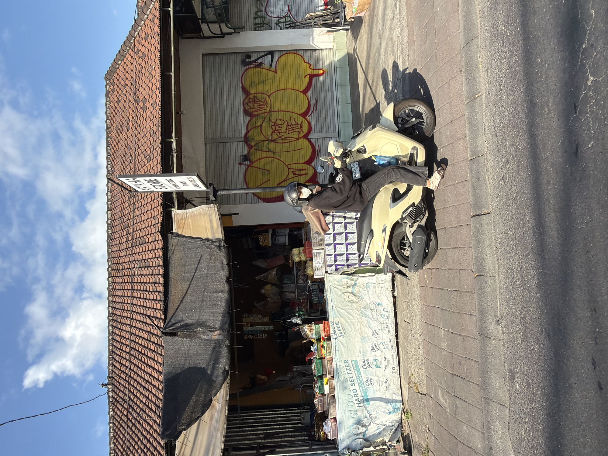 A person wearing a black hoodie, dark pants, a helmet, and a face mask rides a light yellow scooter on a sidewalk in North Kuta, Indonesia. The scooter is positioned in the foreground, facing right. Behind the scooter is a store with a tiled roof, a black awning, and a display of goods. A banner across the front of the store reads "CLOUD SEVEN HARD SELTZER". Above the store entrance, a sign says "ARIKHA STORE COSMETIC DAN AKSESORIS". The building's shutters are decorated with yellow graffiti. The sidewalk is paved with square tiles, and the street is asphalt. The sky is blue with scattered clouds.