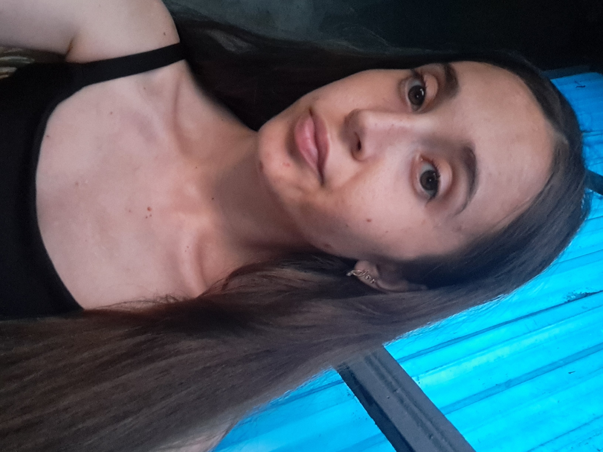 A young woman with long, dark brown hair is pictured from the chest up, looking directly at the camera. She is wearing a black tank top. Her skin has some blemishes. She is wearing a small, ornate earring in her left ear. The background is a blue, corrugated surface, possibly siding or a fence, with some dark vertical elements suggesting a structure. The image appears to be taken outdoors. There is no specific indication of the location within Vypasne, Ukraine.