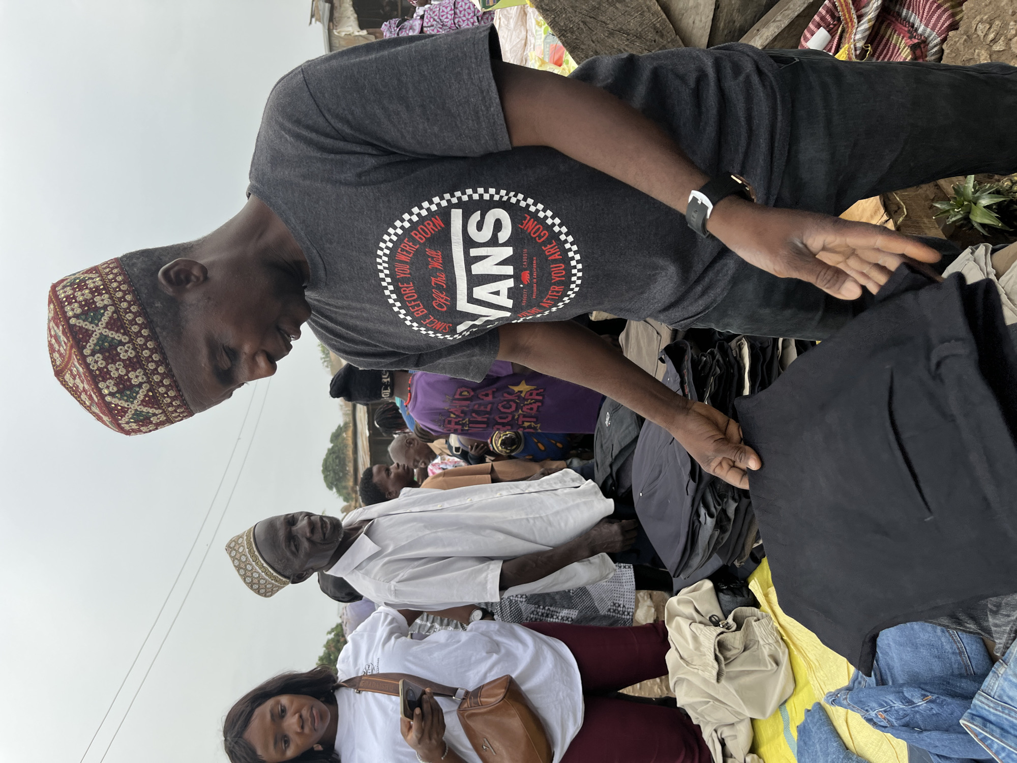 Here's a detailed description of the image:
**Scene:** The image appears to have been taken at an outdoor market in Gura Top, Nigeria. There's a crowded atmosphere with people closely gathered around merchandise.
**People:**
* A man wearing a gray t-shirt with a "Vans Off The Wall" logo and a decorative cap is holding up a black piece of clothing, examining it. He also has a wristwatch.
* Behind him and to the left, another man wearing a similar decorative cap and a white shirt is visible.
* In the foreground, a woman is holding a mobile phone, also wearing a white t-shirt.
**Objects:**
* Piles of clothing are spread out, seemingly for sale. There are jeans, a tan drawstring bag and other garments visible.
* There is a pineapple nearby.
**Actions:**
* The man in the Vans shirt is inspecting a black garment.
* The woman in the foreground is looking at her phone.
**Environment:** The setting is outdoors, likely a marketplace. The lighting appears somewhat overcast.
