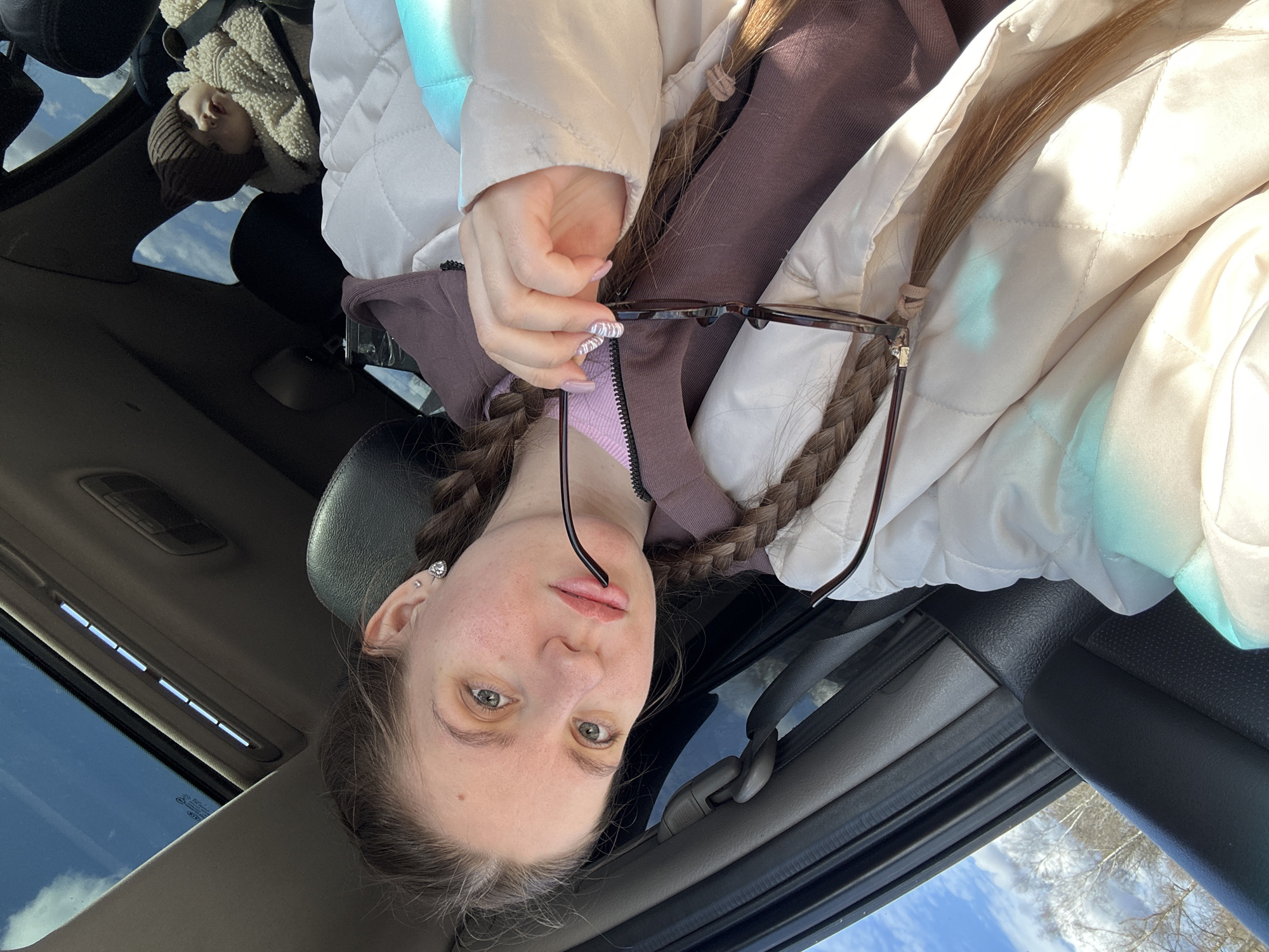 A selfie photograph taken from inside a car with an open sunroof. The photographer, a young woman with green eyes and blonde hair in braids, is wearing a quilted jacket, a purple hooded sweatshirt, and an earring in her left ear. She holds a pair of eyeglasses in her right hand. In the car, in the back seat, is another person wearing a fur coat and a brown beanie. The car's interior is visible, including the sunroof, seats, and window frames. Outside, the sky is blue with some clouds and bare trees are visible. It's unknown if the image was taken in Klin, Russia.