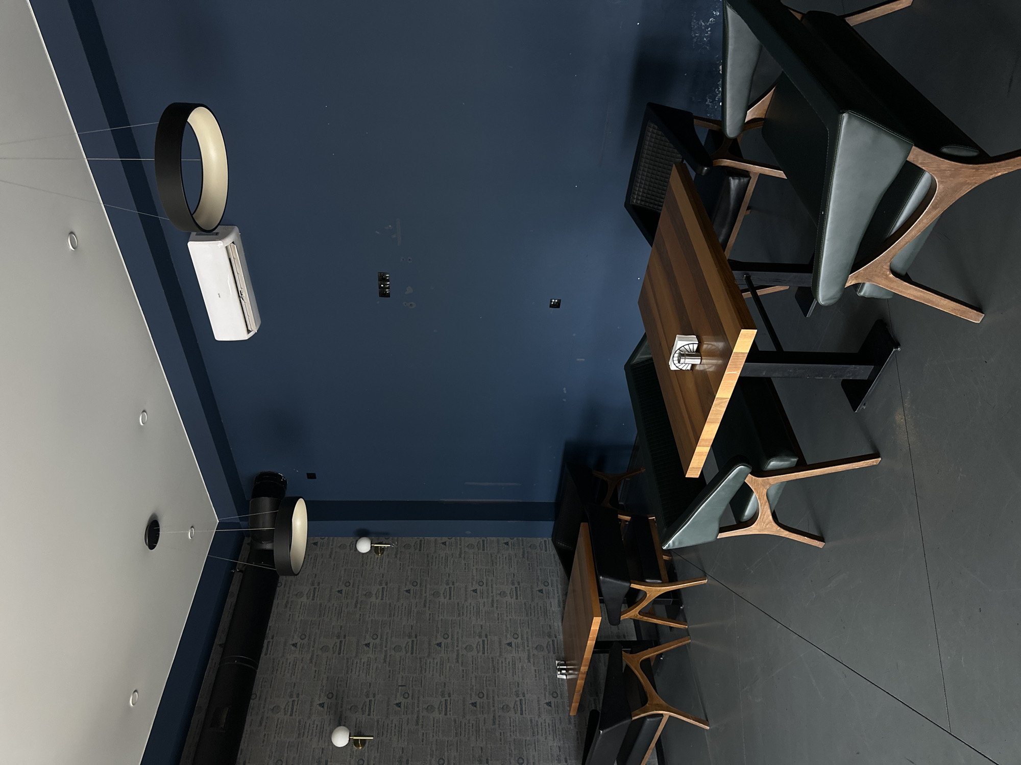 A bird's-eye view shows the interior of a contemporary establishment in Marneuli, Georgia. The ceiling is partitioned into a white section, a deep blue segment, and a patterned gray portion. A ring-shaped lamp hangs suspended near an air conditioning unit. Below, wooden tables are arranged alongside gray chairs with wooden frames. The floor alternates between patterned gray and a solid dark shade. A dark, vertical pipe runs along the wall from floor to ceiling. The scene is devoid of people.