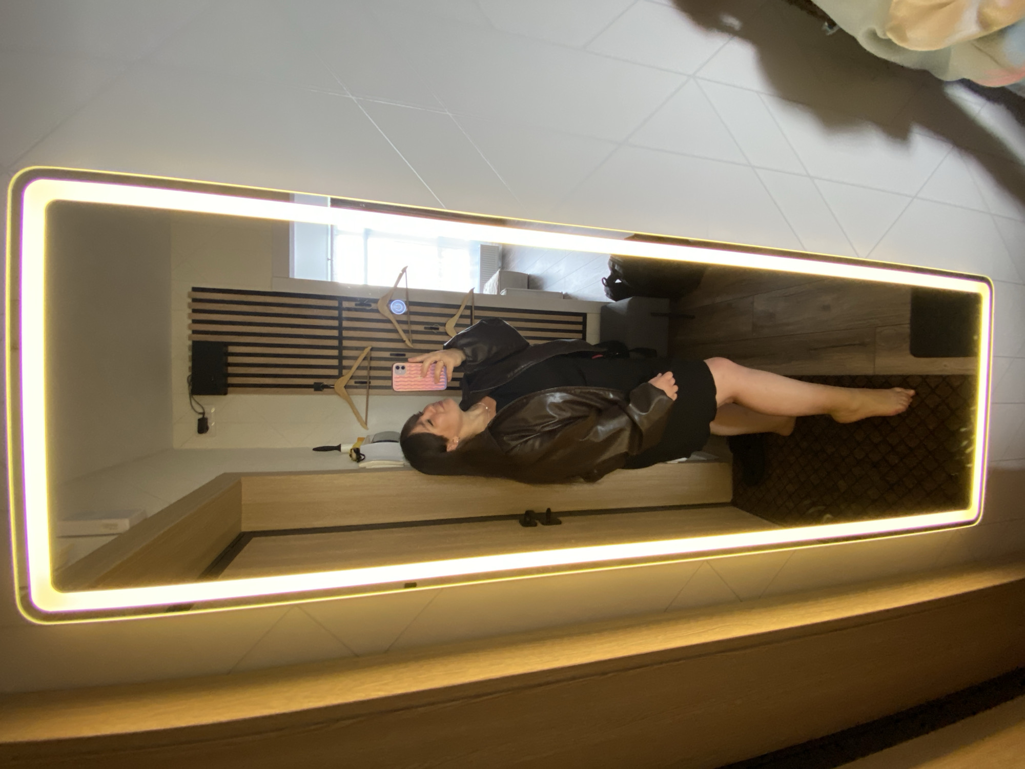 A woman is depicted taking a mirror selfie in a modern room, likely in Kazan, Russia. The image captures her reflection in a long, rectangular mirror that is mounted vertically on a wall and features a vibrant, warm yellow-white illuminated border. The woman is lying horizontally on her side on what appears to be a bed or a low bench. She is propped up, holding a pink smartphone with a patterned case in her right hand, positioned to capture her reflection.
She is dressed in a dark top or dress and a dark leather jacket, with her bare legs extended. Her bare feet rest on a dark, geometrically patterned rug or floor covering.
The room's decor, visible in the mirror's reflection, includes light-colored walls and a distinctive wooden slatted panel, possibly a headboard or accent wall. A window is visible in the background, along with a small black device, potentially a speaker, mounted on the slatted panel, and some wooden hangers or decorative elements. A light-colored wooden surface, likely part of the bed frame or a low console, runs along the base of the mirror. The overall ambiance is contemporary and comfortable.