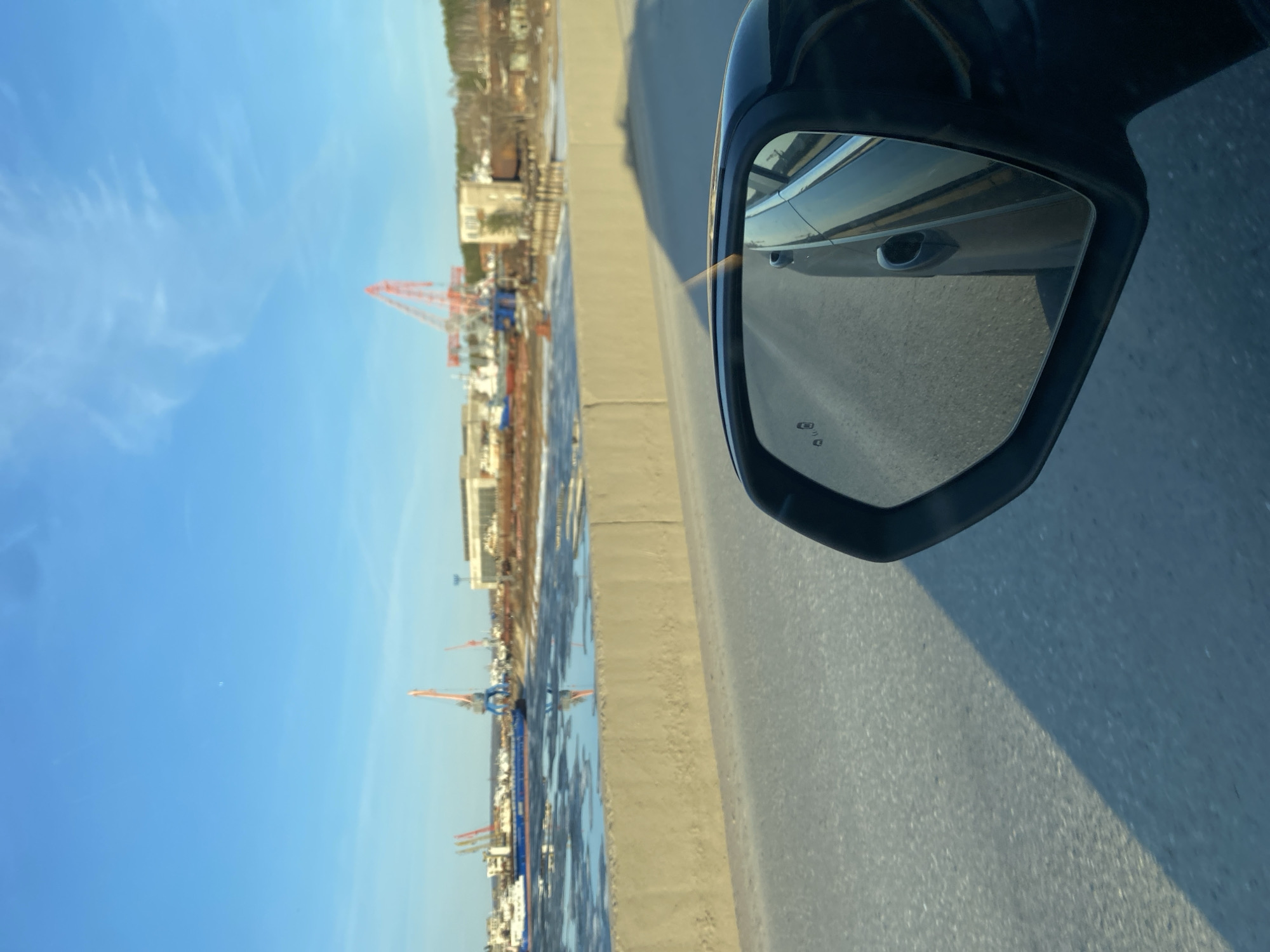 The image, taken from a moving vehicle, captures a bright daytime scene along a waterway in Chaikovsky, Russia.
In the foreground, the black side mirror of a car occupies the right side of the frame. Its reflective surface clearly shows a portion of the car's body, including a silver door handle, and the paved road surface alongside the vehicle. Two small icons, indicative of blind spot monitoring systems (resembling cars with arrows), are visible in the lower left corner of the mirror's reflection.
To the left of the road, a body of water, likely the Kama River, stretches into the distance. Patches of ice or slush are visible on the water's surface, suggesting cold weather. Along the far bank of the waterway, a bustling industrial or port area is visible. Numerous large cranes, some red and others orange and blue, stand prominently against the sky, indicating active construction or cargo handling. Various buildings and structures line the distant shore.
The sky above is a clear, vibrant blue with a few wispy white clouds scattered across it. The lighting is bright, consistent with morning or afternoon sun, and the overall atmosphere appears calm. No specific text is legible beyond the aforementioned mirror icons. The scene depicts a typical industrial landscape alongside a river in a cold climate.