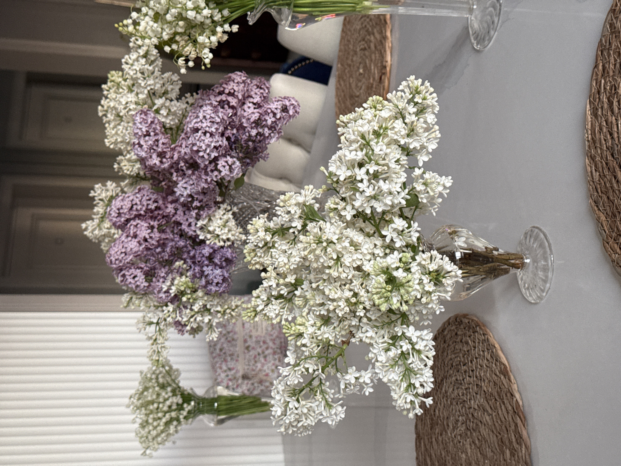 The image shows a table arrangement featuring various flowers in vases, including white lilacs, purple lilacs, and lily of the valley. The vases are placed on a light grey tabletop, with woven placemats underneath some of them. The background reveals glimpses of a room with blinds and other decorative elements, suggesting an interior setting, perhaps in Moscow, Russia, though this cannot be confirmed from the image alone. There are no people visible, and the scene focuses on the floral arrangement as the main subject.
