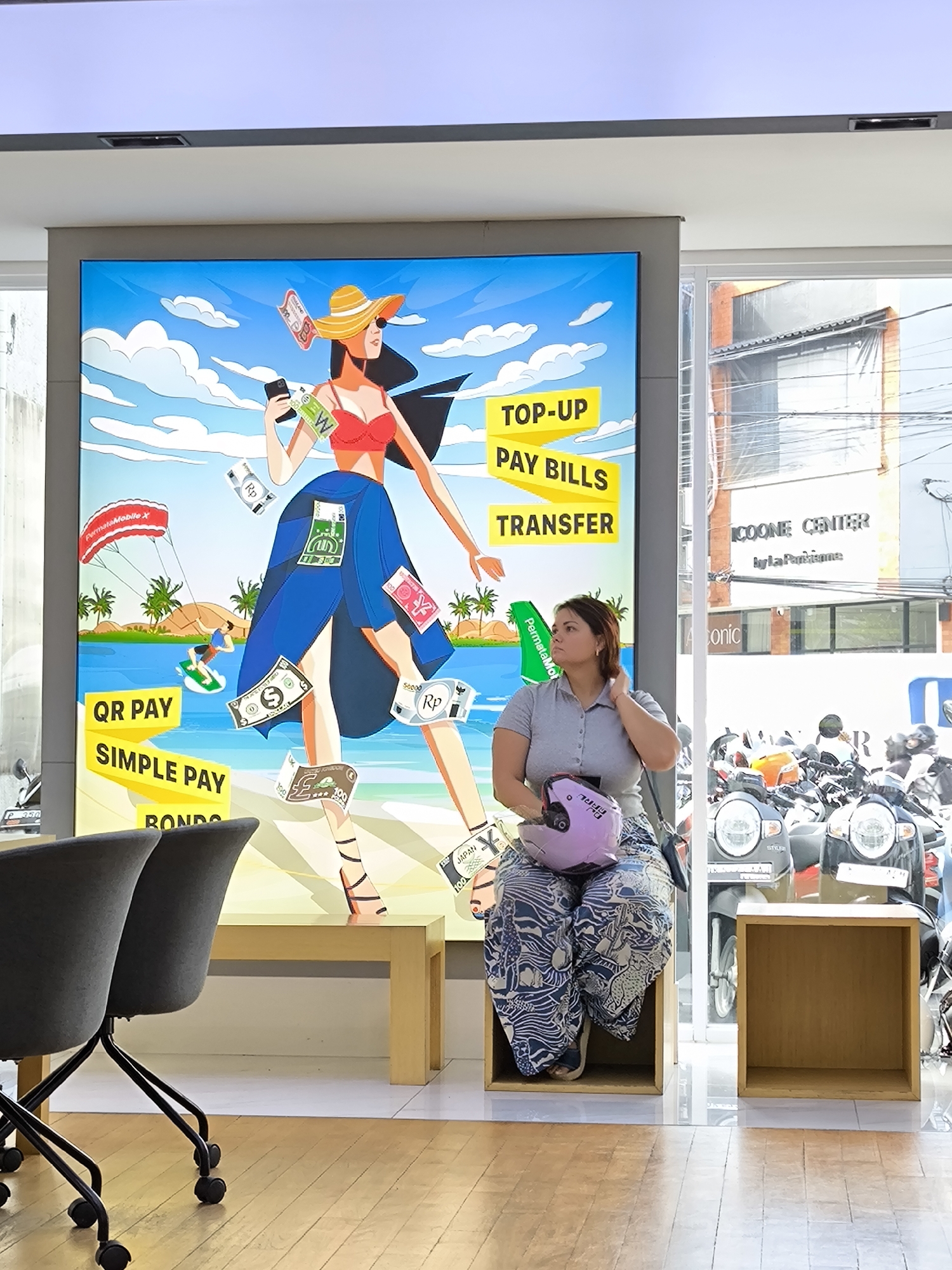The photograph depicts a young woman sitting on a wooden box in what appears to be a business setting, possibly a bank or financial institution in North Kuta, Indonesia. She is light-skinned with auburn hair, wearing a grey polo shirt and patterned blue and white pants. She's holding a purple helmet with a white design on it, and her left hand is raised to her head.
Behind her is a large, brightly colored advertisement for PermataMobile X, featuring a stylized woman in a bikini top and blue skirt, seemingly surrounded by money from various countries. The text in the ad highlights services like "Top-Up, Pay Bills, Transfer, QR Pay, Simple Pay, and Bonds."
Through the glass window to the right of the woman, a street scene is visible, with several motorcycles parked outside. Above the bikes, a building is partly visible with signage that reads "ICOONE CENTER by la Parisienne."
In the foreground to the left are two grey office chairs, indicating the business environment. The flooring is light wood. To the right, another wooden box stands, similar to the one the woman is sitting on. The image appears well-lit, suggesting an interior space.