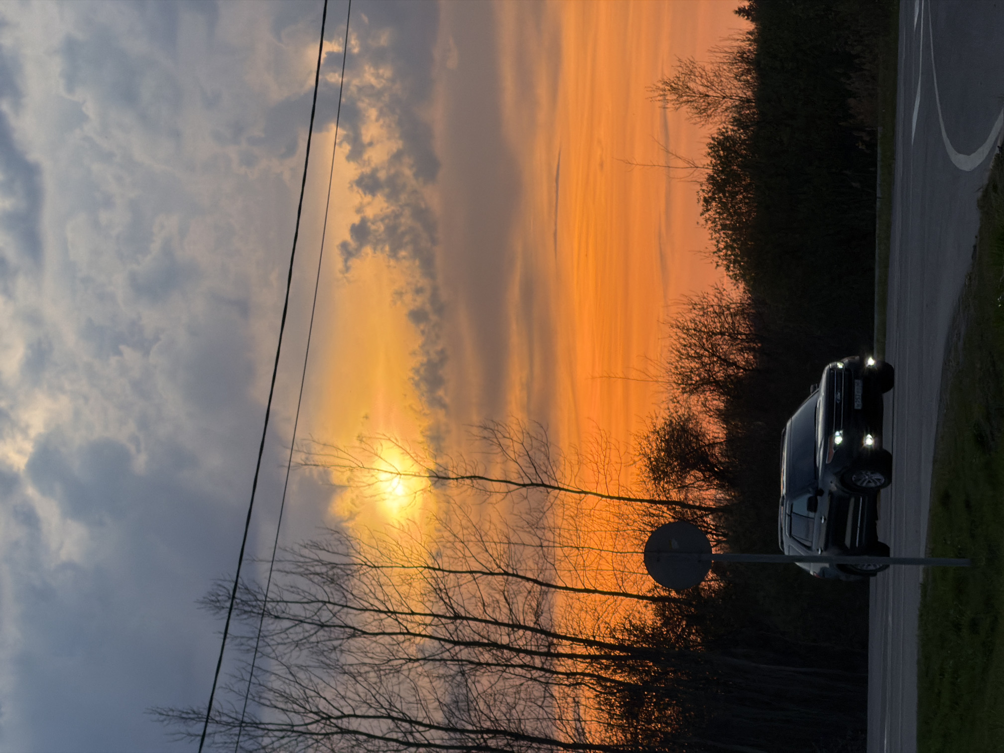 The image shows a sunset in Ostrov, Russia. The sky is split between dark clouds on the left and orange and yellow sunset on the right. Silhouetted bare trees are in the foreground, with power lines running across the sky. A car with headlights on is parked on the right side of the image, next to a road. A round traffic sign is near the car.