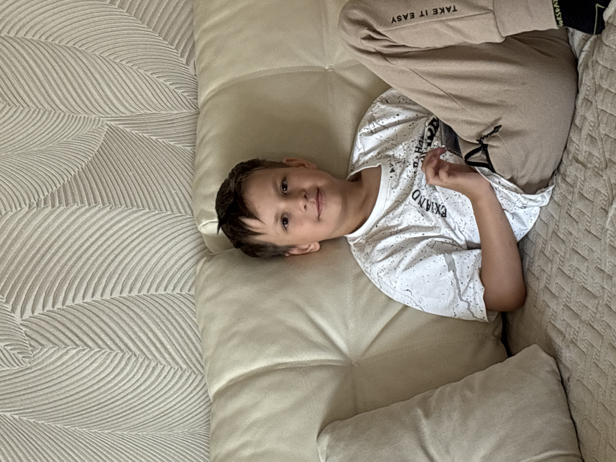 A boy lies on a beige, quilted sofa in an indoor setting. He is wearing a white t-shirt with black lettering, and light brown sweatpants with "TAKE IT EASY" printed on the right leg. The sofa is patterned with a wave-like design on the left side. The exact location is not discernible from the image but the city of Chaikovsky, Russia was provided.