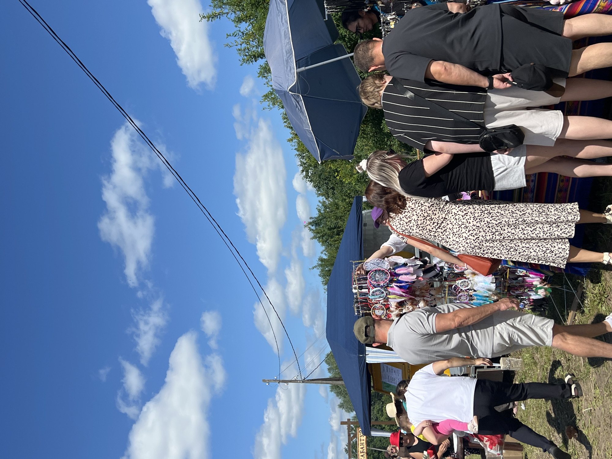 A group of people are gathered around a market stall displaying dreamcatchers on a sunny day in Kalinino, Russia. A blue tent is set up behind the stall, and trees form a backdrop against a bright blue sky with scattered white clouds. Several individuals are examining the merchandise, with one person holding up a dreamcatcher. The ground is covered in dry grass and dirt. Electrical wires are visible stretching across the sky, connected to a wooden utility pole.