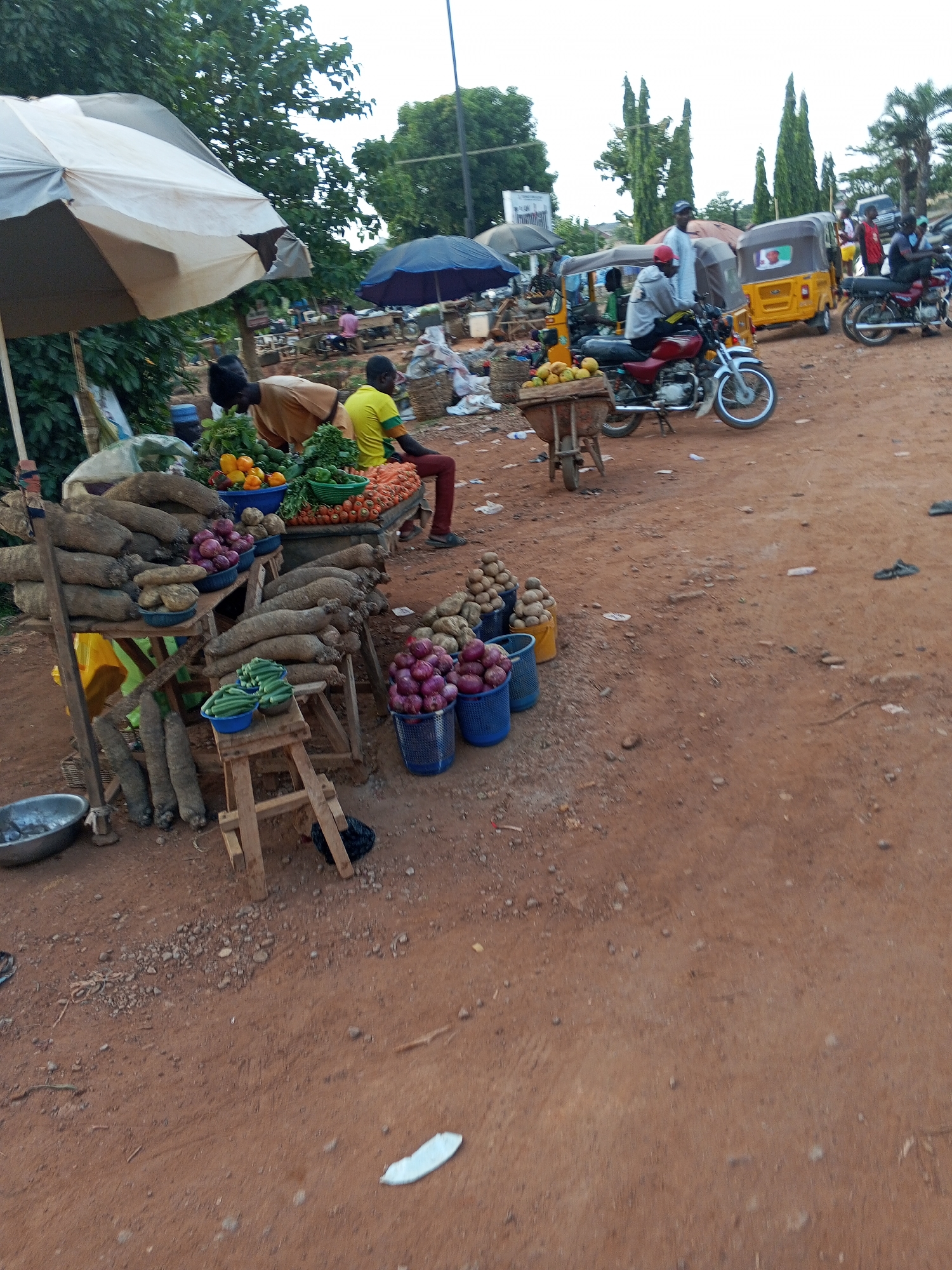 The image shows a market scene in Galadimawa Village, Nigeria. Stalls display yams, onions, green vegetables, and oranges. Some vendors sit by their produce. Motorbikes and a tricycle are parked nearby. The ground is reddish-brown dirt. People are present, some wearing brightly colored shirts. One man sits on the edge of a produce table. Several motorcycles are present. Overhead, there is an off-white canopy tent over a produce display.
