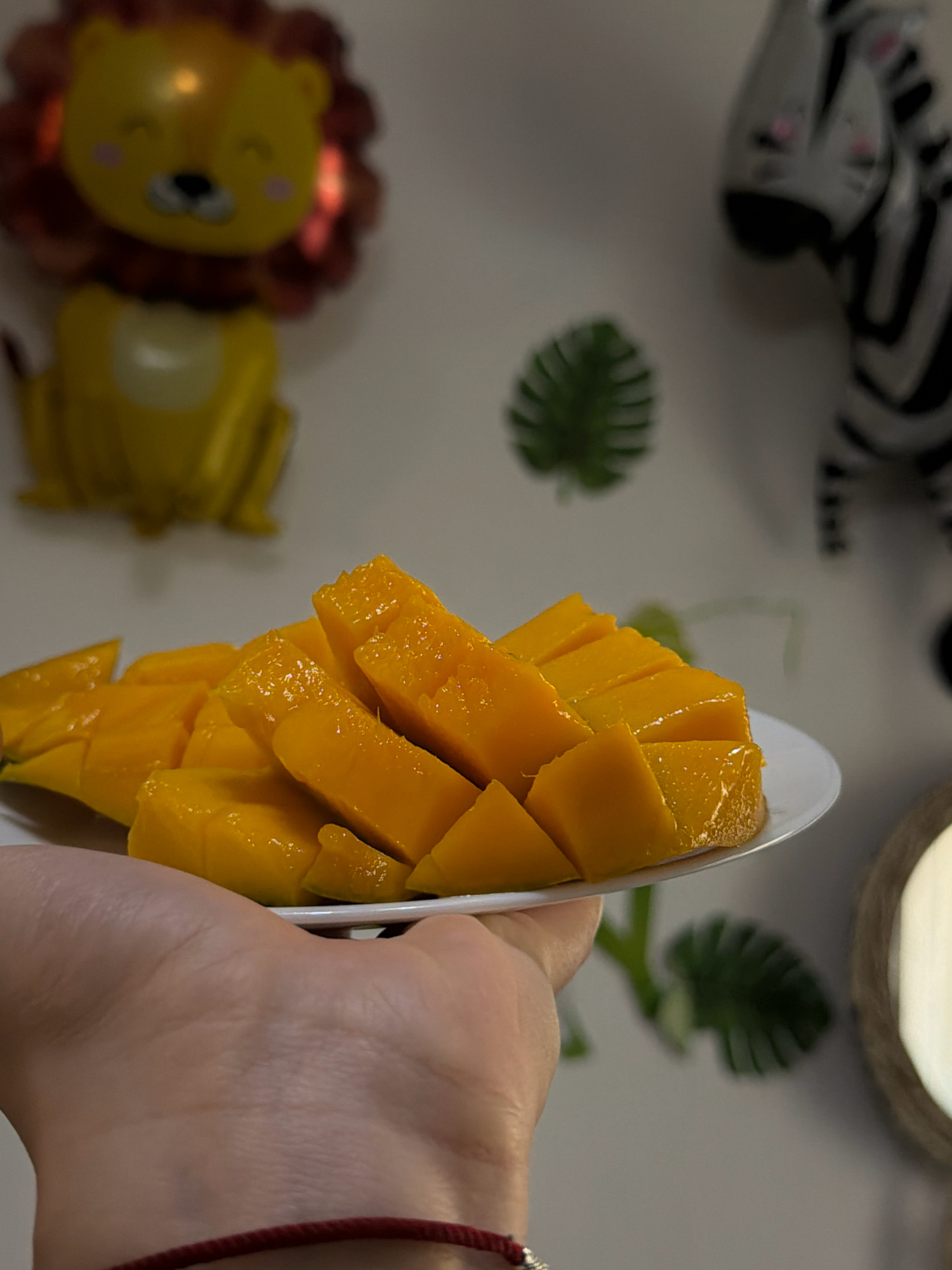 A hand is holding a white plate filled with sliced mangoes. In the background, out of focus, are decorations that appear to be for a children's party, including a lion-shaped balloon and a zebra-shaped decoration. There are also some artificial green leaves visible. The overall scene suggests a casual, domestic setting, possibly a home or a small gathering space. There is no identifiable information in the image that places it in Sharm El Sheikh, Egypt.