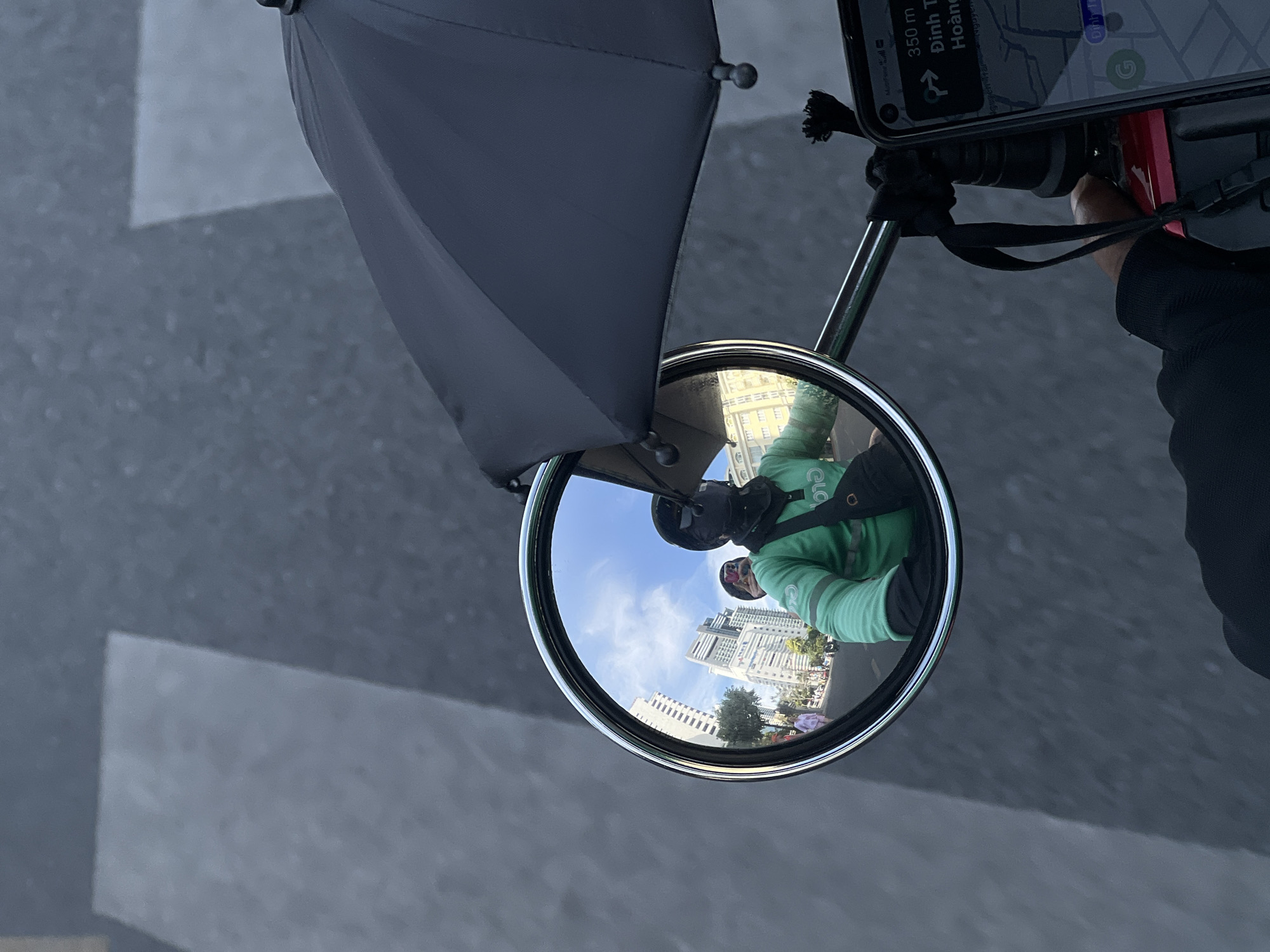 The image is a first-person perspective from what appears to be a motorbike or scooter, equipped with a side mirror, a phone mounted to the handlebars, and an umbrella. The reflection in the side mirror shows the rider, wearing a helmet and a green shirt with "Grab" branding, as well as the background cityscape, featuring buildings and a blue sky. A navigation app is visible on the phone screen, displaying "350 m Dinh T Hoang". The setting is Nha Trang Ward, Vietnam, indicated by the navigation app. The background is blurred. The focus is on the mirror reflection.