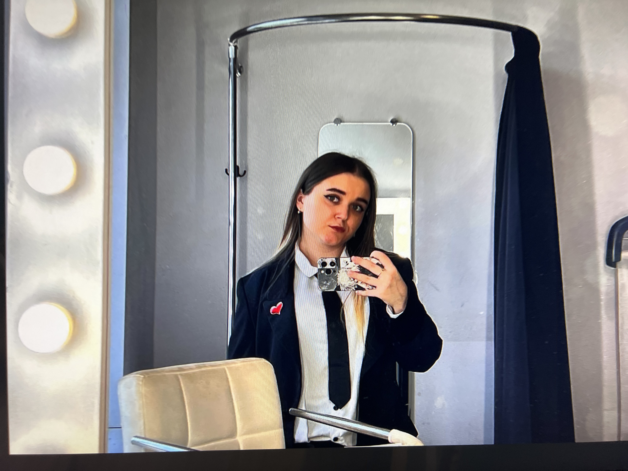 This media file is a photograph of a screen displaying a mirror selfie of a young woman, likely taken in Pushchino, Russia. The woman, with long dark hair, is positioned centrally, looking directly into the camera reflected in a large mirror. She wears a black blazer, a white collared shirt, and a black tie. A small red heart-shaped pin is attached to the lapel of her blazer. She is holding a smartphone with a patterned case to capture the image.
The setting appears to be an indoor space, possibly a dressing room or vanity area, characterized by several elements. To the left of the main mirror, a vertical panel with two round, illuminated lights suggests a vanity mirror setup. Behind her, a smaller rectangular mirror is visible, reflecting more of the room. A metallic, curved bar, possibly part of a clothes rack or a curtain rail, is seen above the smaller mirror, and a dark curtain hangs to the right. A light-colored, upholstered chair is partially visible in the foreground, below the main mirror. The walls are a plain, light grey. The woman's expression is neutral, and her makeup includes eyeliner and lipstick. There is no visible text in the image.