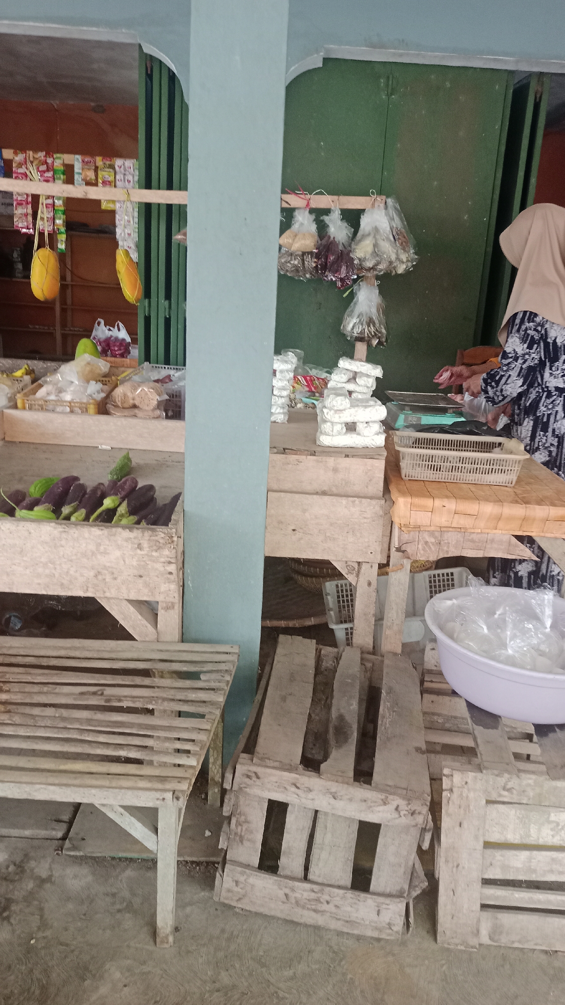 The image depicts an outdoor market stall in Karyamandala, Indonesia. The stall is partially covered by a turquoise-colored beam and a green corrugated metal wall. In the foreground, a wooden bench is visible on the left. A wooden crate filled with purple eggplants and a few green gourds is also present. To the right, several wooden crates and pallets are stacked.
Further back, various food items are displayed. Packets of snacks and yellow fruits hang from a wooden beam on the left. A stack of white, rectangular food items, likely tempeh, is prominent in the center. Behind the tempeh, a person wearing a beige hijab and a patterned dress is seen interacting with a scale and plastic bags, suggesting they are a vendor. Bags of dried goods, such as grains and spices, are suspended from another wooden beam. A white plastic bowl filled with small, white objects, possibly crackers or snacks, is on the right. The ground is concrete. The overall impression is a functional, albeit somewhat rustic, market setting.