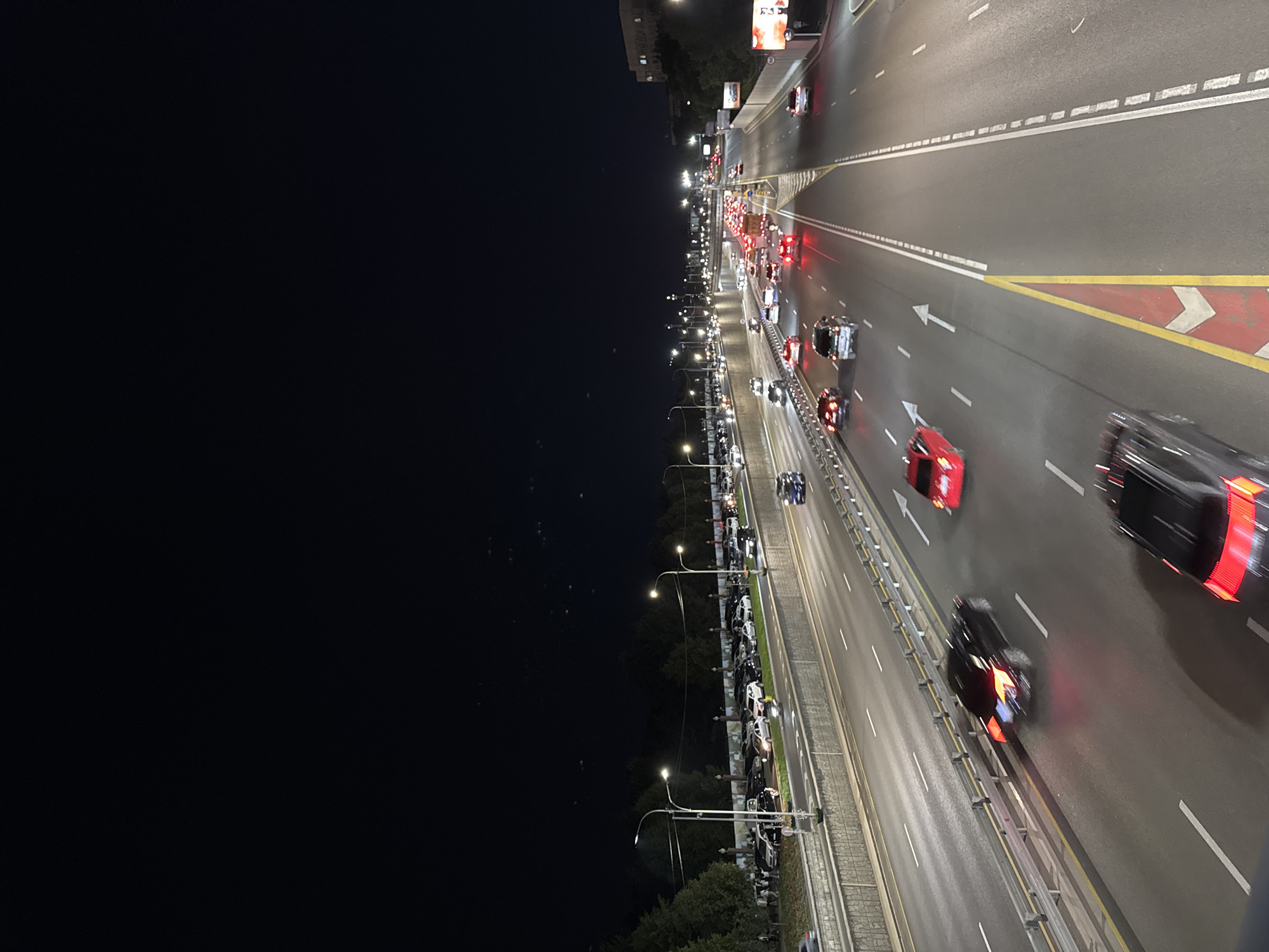 The image depicts a night-time scene of a multi-lane highway with moderate traffic in Almaty, Kazakhstan. The highway appears to be elevated, with a retaining wall and a sidewalk bordering the lanes. On the sidewalk, several parked cars and a few pedestrians are visible. The highway itself features numerous vehicles, many with their taillights illuminated, indicating ongoing traffic. The road surface is paved, and lane markings are clearly visible. Streetlights illuminate the highway, casting a glow on the vehicles and the surrounding environment. Trees and buildings line the sides of the highway, and the sky above is dark, suggesting it is nighttime. The overall impression is that of a busy urban road at night.