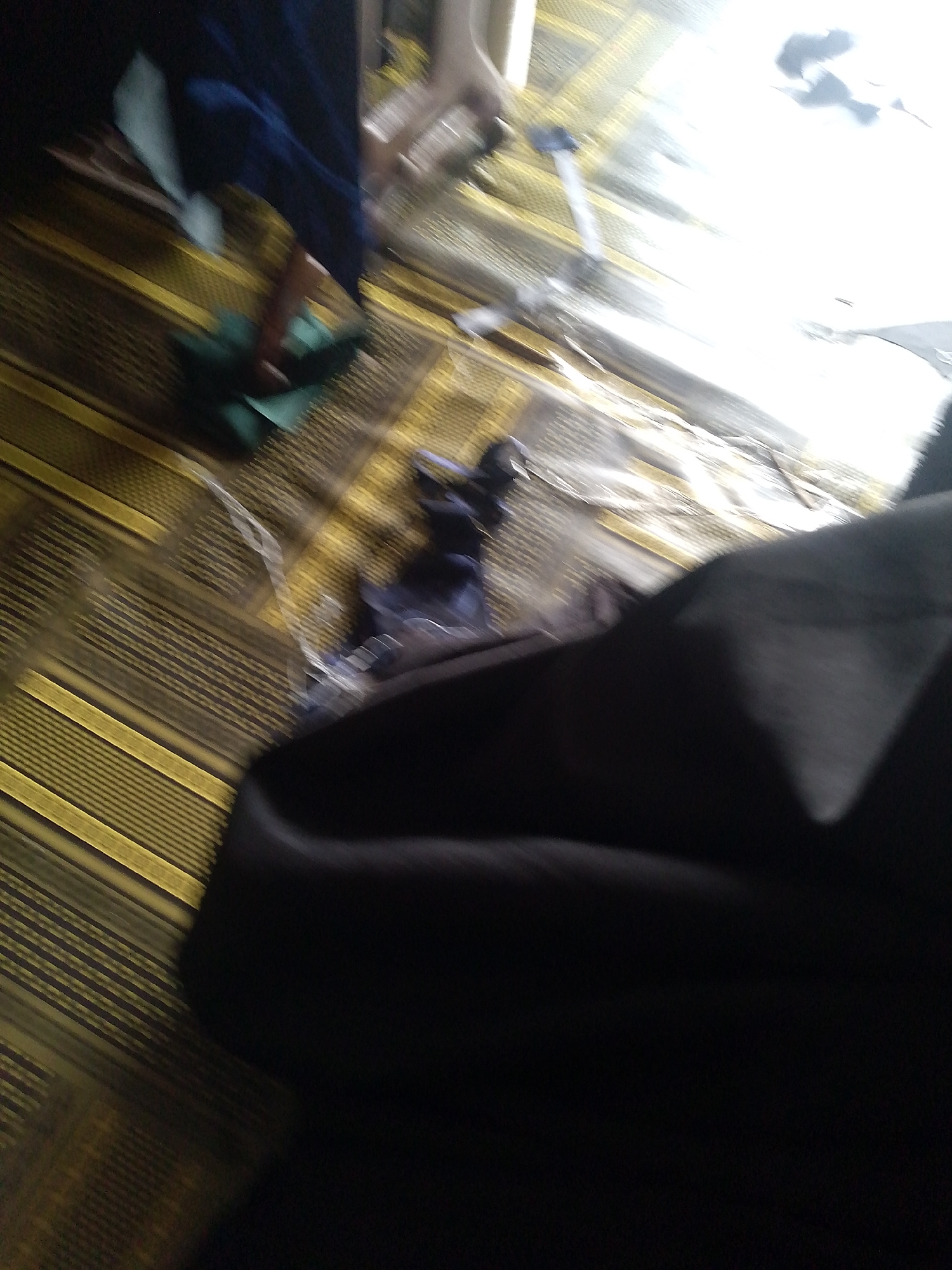In the city of Jauro Yinu, Nigeria, a dimly lit room is captured with a blurred motion. The floor is covered with a yellow and brown striped rug. In the lower left corner, a dark blue cloth lies draped over a green cloth. Several other dark-colored fabrics are scattered across the rug, some appearing crumpled. In the upper portion of the image, a bright white light source causes a significant glare and obscures details. The overall scene suggests a domestic or storage space with everyday items present.