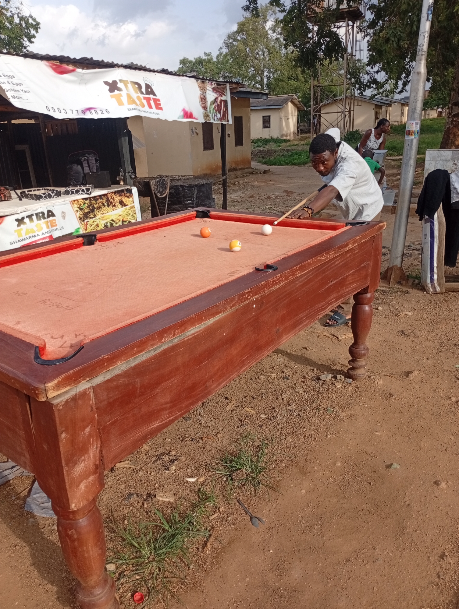 In Rafin Par, Nigeria, a man plays billiards outdoors in front of a food stall. The man, wearing a white t-shirt and brown sandals, is poised to strike a white billiard ball with a cue. The billiard table, a rustic wooden structure with a reddish felt surface, is positioned on uneven, dusty ground. Several billiard balls are scattered on the table, including an orange one and a yellow one. Behind the table, a banner for "XTRA TASTE SHAWARMA AND GRILLS" is visible, indicating the presence of a food vendor. The environment appears to be a developing area with simple buildings, trees, and a water tower in the background. Another person, a woman, is visible in the middle ground, carrying a bucket. The sky is partly cloudy.