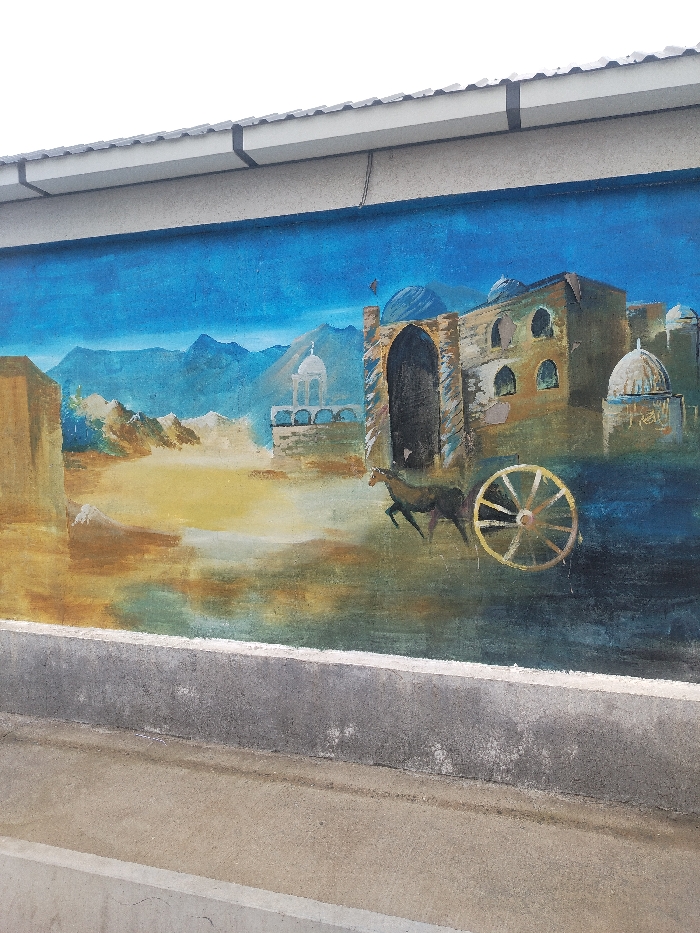 A painted mural is visible on a concrete wall in the city of Toytepa, Uzbekistan. The upper section of the wall features a white gutter system with black vertical supports, positioned beneath an overcast sky. The mural itself occupies the central area, depicting a stylized landscape. The background of the mural consists of layered blue mountains beneath a gradient blue sky. The midground transitions into an arid, yellow-brown landscape with a reflective, darker surface at its base. Architectural elements within the mural include a large, multi-arched, domed building on the right, and a smaller domed structure with supporting arches in the central-left. A rectangular, light-brown building is depicted on the far left. A dark-colored horse in mid-stride and a large, spoked wooden wheel are prominently featured in the lower-right portion of the mural. A small, red, flag-like object is visible above the large building on the right. Below the mural, the wall transitions to a grey concrete base, beneath which a light brown, flat ground surface is partially visible. The mural exhibits some minor discoloration and surface wear.