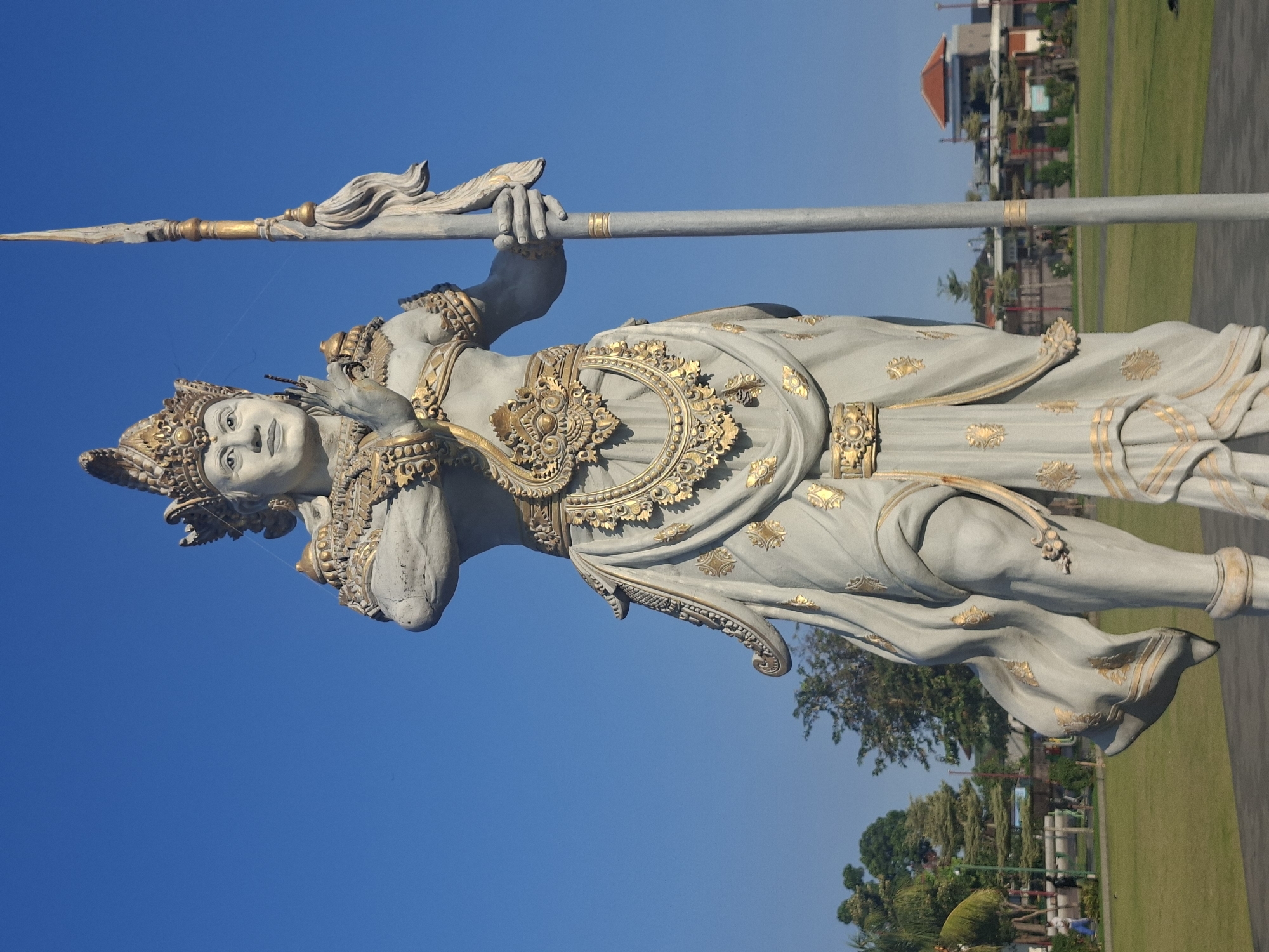 A low-angle, full-body shot captures a large, grey stone statue of a warrior deity against a clear, bright blue sky. The statue is adorned with intricate gold embellishments on its crown, jewelry, and clothing. It holds a long, grey spear vertically in its right hand. The statue stands on a dark grey base in an open, grassy area. In the background, lush green trees and a building with a red roof are visible. The overall scene suggests a public space or monument in Gianyar, Indonesia, given the style of the statue, which is typical of Balinese art and architecture.