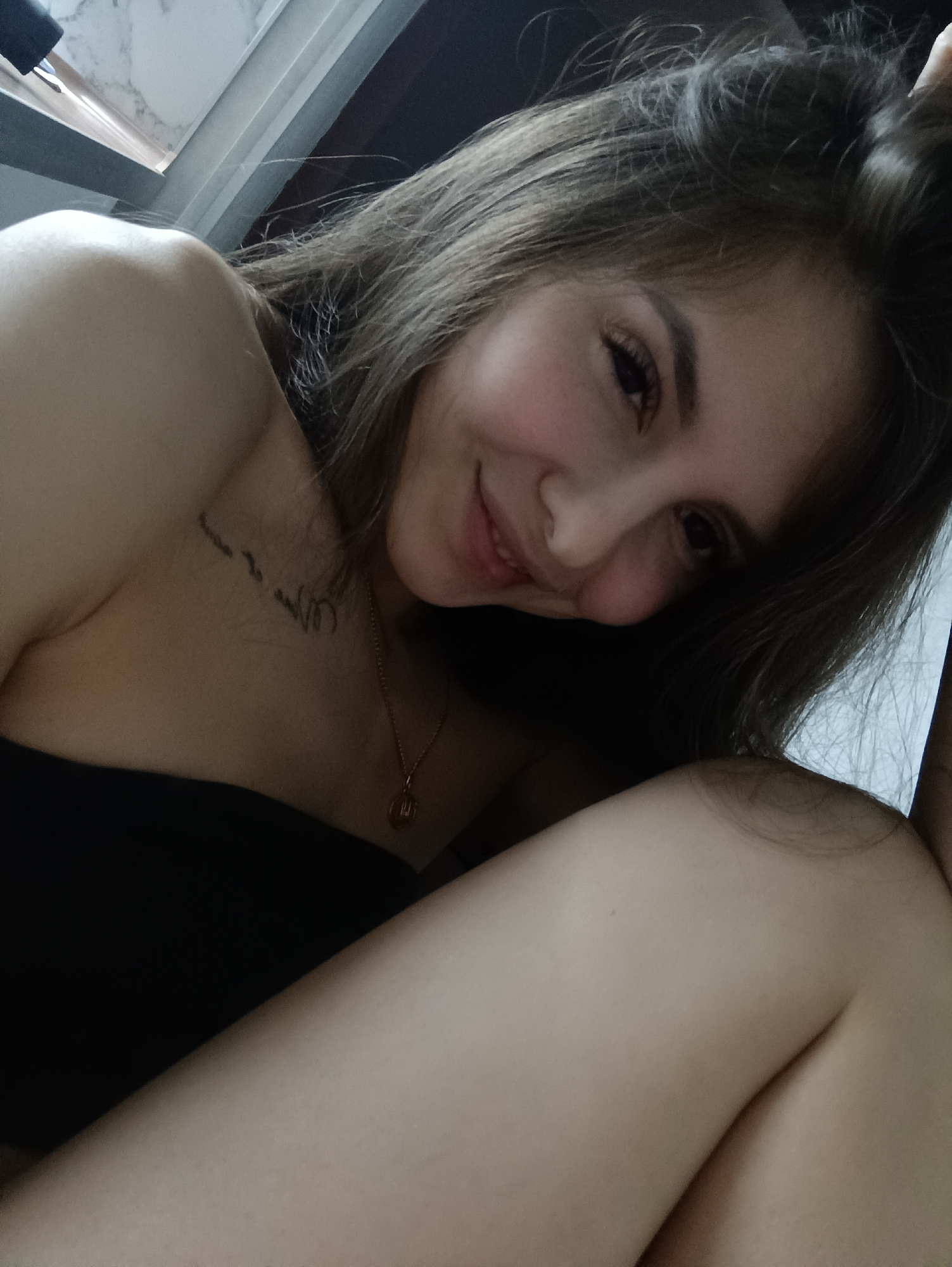 The image shows a young woman with brown hair smiling at the camera. She's wearing a black strapless top and a gold necklace with a small pendant. There is a tattoo visible on her upper chest. The background appears to be indoors, with a glimpse of a window frame or countertop in the upper left corner. The exact location in Chaikovsky, Russia, cannot be determined from the image.