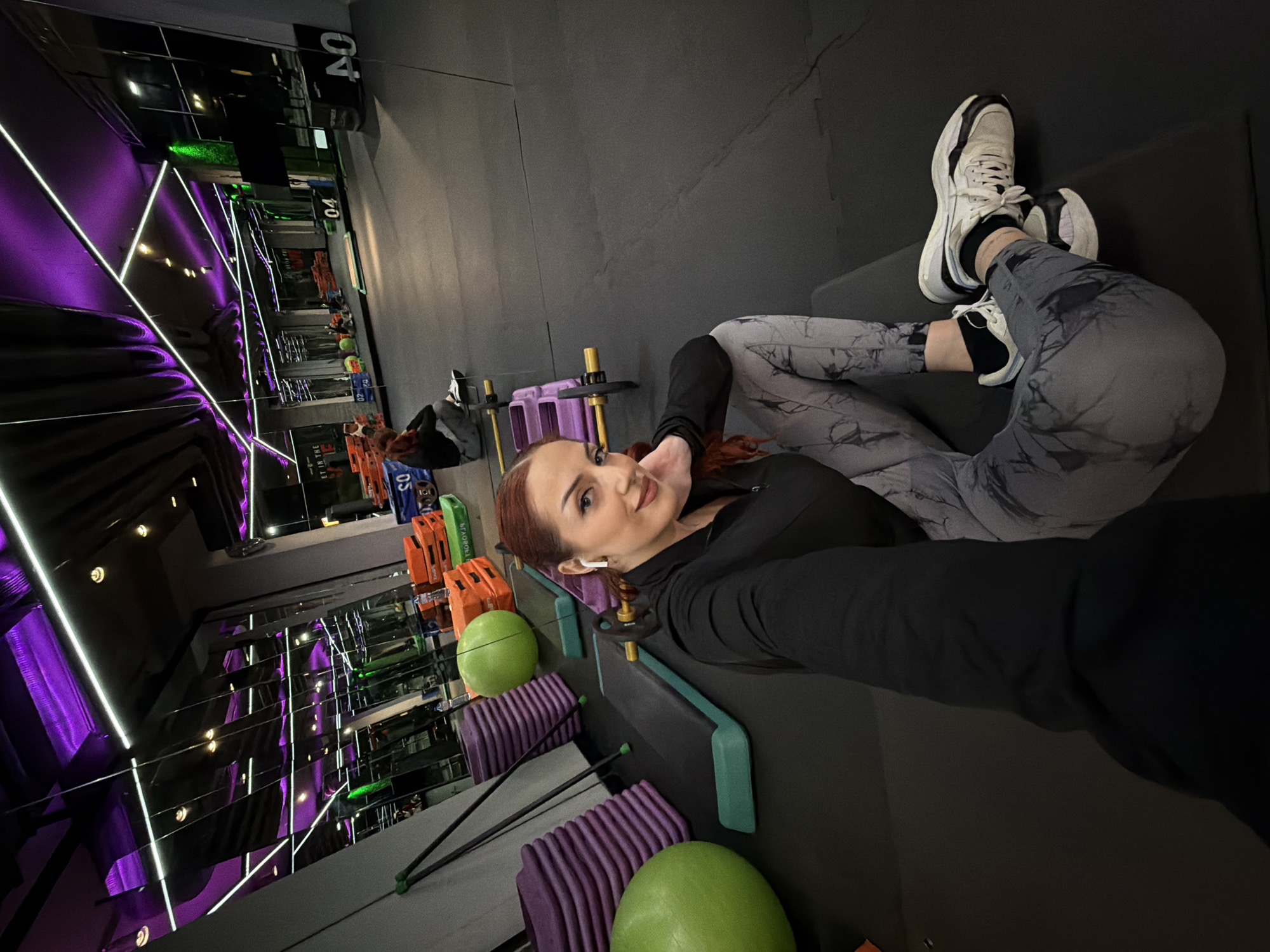 A woman is lying on an exercise bench at a gym. She wears grey marble leggings and a black long-sleeved shirt. Her shoes are white with black accents, and she wears black socks. She appears to be holding the camera in her extended right hand to take a selfie. She has red hair and is wearing wireless earbuds. The gym features equipment and purple LED lighting, likely in Sofia, Bulgaria.