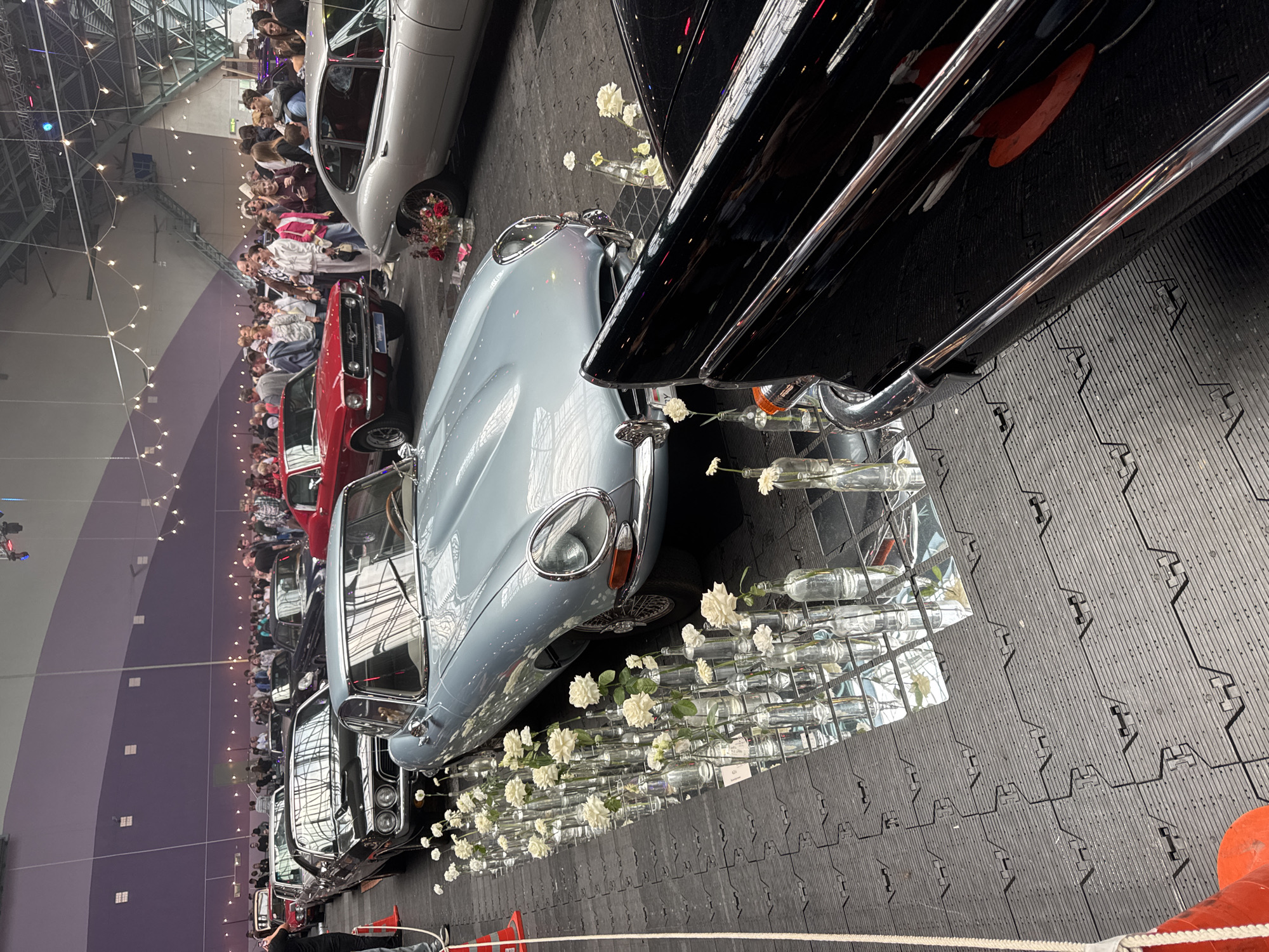 The image depicts an indoor exhibition of classic cars. In the foreground, a light blue vintage Jaguar E-Type is prominently displayed, with a row of glass bottles containing white roses placed along its side on a mirrored surface. To the right of the E-Type, the polished black rear section of another classic car, possibly a Cadillac, is visible. In the background, various other vintage cars, including a red Ford Mustang, are parked in rows, and a crowd of people are milling around. The environment appears to be a large hall with a high ceiling, strung with decorative lights. The floor is covered with black interlocking mats. The exhibition is likely taking place in Minsk, Belarus.