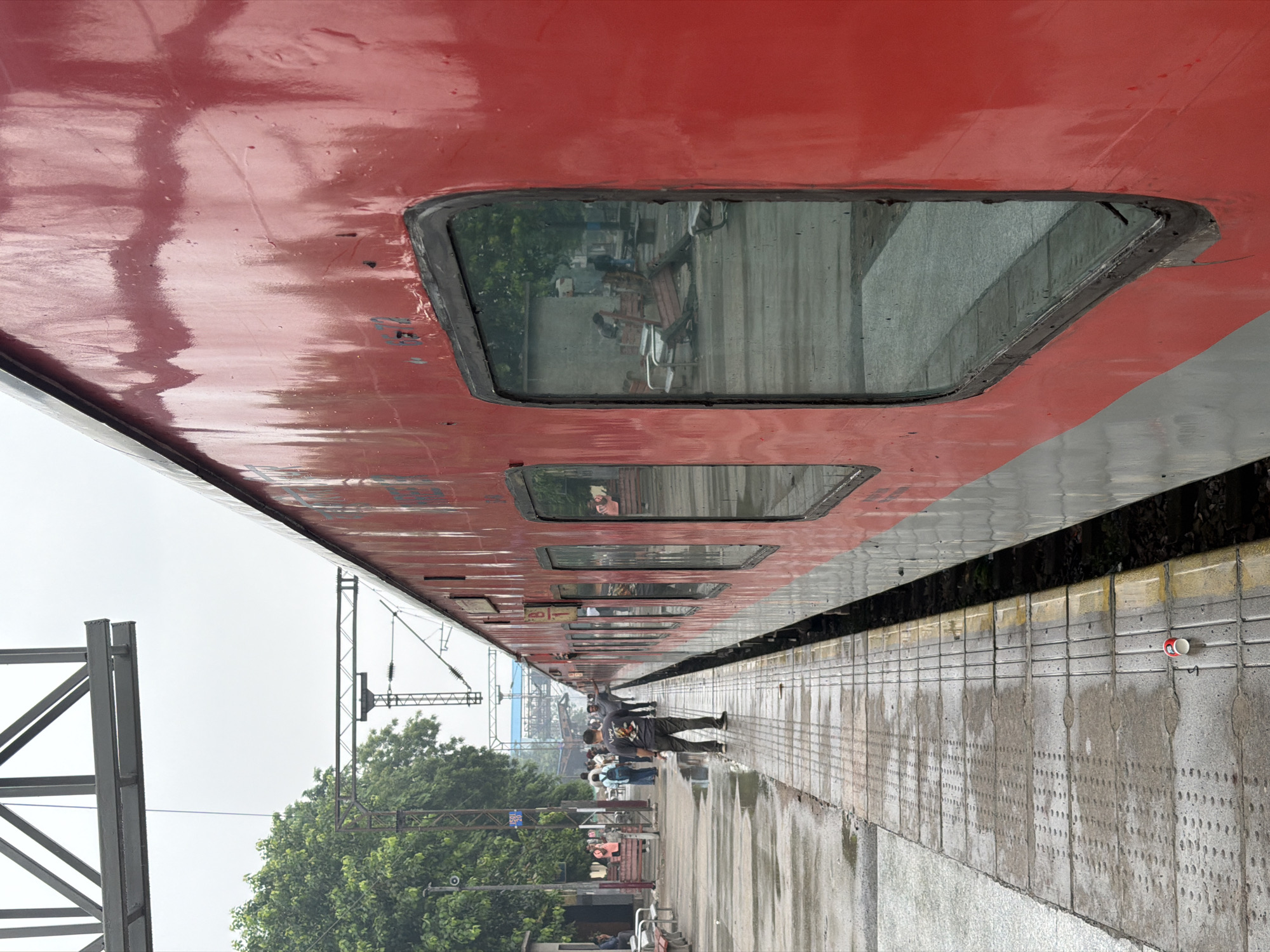 The image depicts a red and grey train at a train station platform in Ballabgarh, India. The platform is wet, with visible puddles and water stains on the concrete tiles, suggesting recent rain. Several people are visible on the platform. In the foreground, a man wearing a dark t-shirt and pants stands facing away from the camera, looking towards the train. Further down the platform, a group of people are congregated, some appear to be waiting or disembarking. Trees line the left side of the platform, and overhead electrical lines for the train are visible. The windows of the train reflect the surrounding environment, showing glimpses of structures and foliage. A discarded red and white disposable cup lies on the platform near the tracks. The sky is overcast.