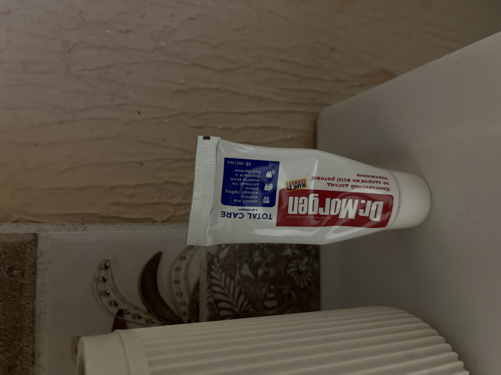 A tube of Dr. Morgen Total Care toothpaste is positioned on a surface, with a portion of a white textured object to its left and a wall with textured wallpaper behind it. The toothpaste tube is white with red and blue lettering. The red lettering clearly displays "Dr. Morgen," and the blue lettering indicates "TOTAL CARE." Some smaller text is visible in both Ukrainian and English, specifying product details. A label reads "MADE IN GERMANY." The background wall has a roughly textured pattern in shades of beige and brown. The white object to the left has a fluted or ribbed surface. The overall scene appears to be indoors, on a shelf or countertop. The image does not provide any information that could be used to determine its location within the city of Kalynivka, Ukraine, or any other specific geographic location.