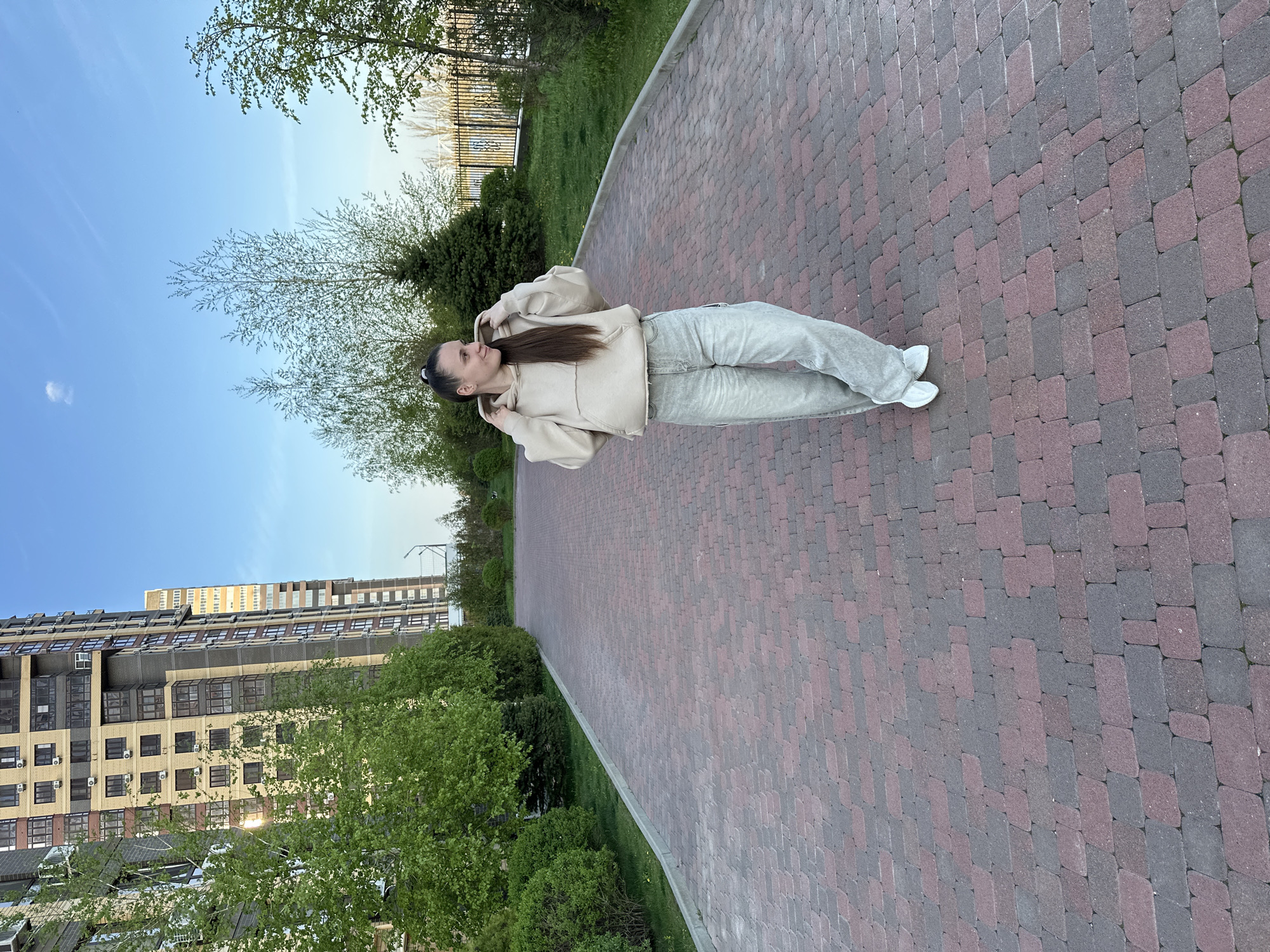 The image shows a woman standing on a brick paved path. She is wearing a beige hoodie, grey sweatpants, and white sneakers. The path is lined with green trees and bushes on both sides. In the background, there is a tall residential building on one side and a blue sky with some trees on the other. The location is Rostov-on-Don, Russia.