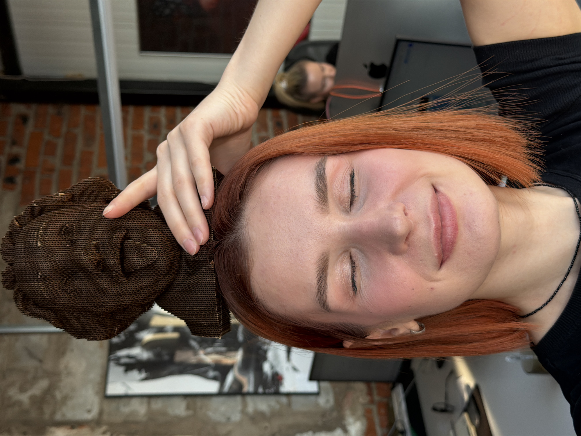 This image depicts a young adult female inside an office or creative workspace located in Chaikovsky, Russia. She has short, straight, reddish-orange hair and her eyes are closed, with lips subtly upturned. Her left hand is positioned on top of her head, supporting a small, dark brown bust made from layered corrugated cardboard. The bust features a stylized human face with an open mouth and an extended tongue, strongly resembling the iconic photograph of Albert Einstein. The female subject wears a black crew-neck shirt and a black beaded necklace. The background reveals a brick wall on the left with a framed, dark-toned artwork. To the right, a window or glass partition is visible. In the blurred mid-ground, a second individual with blonde hair, styled in a ponytail, is seated at a desk, focused on a computer monitor displaying an Apple logo.
