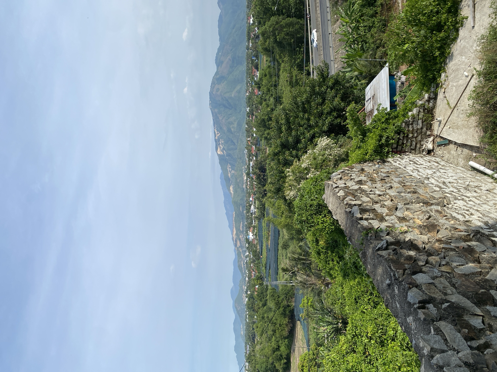 The image depicts a lush, green landscape with a stone wall in the foreground. Beyond the wall, dense foliage gives way to a town nestled among rolling hills and mountains. A highway runs through the scene, with a white car visible. The sky is a pale blue with scattered clouds. The location is identified as Tay Nha Trang Ward, Vietnam. The scene is tranquil and presents a harmonious blend of nature and human habitation.
