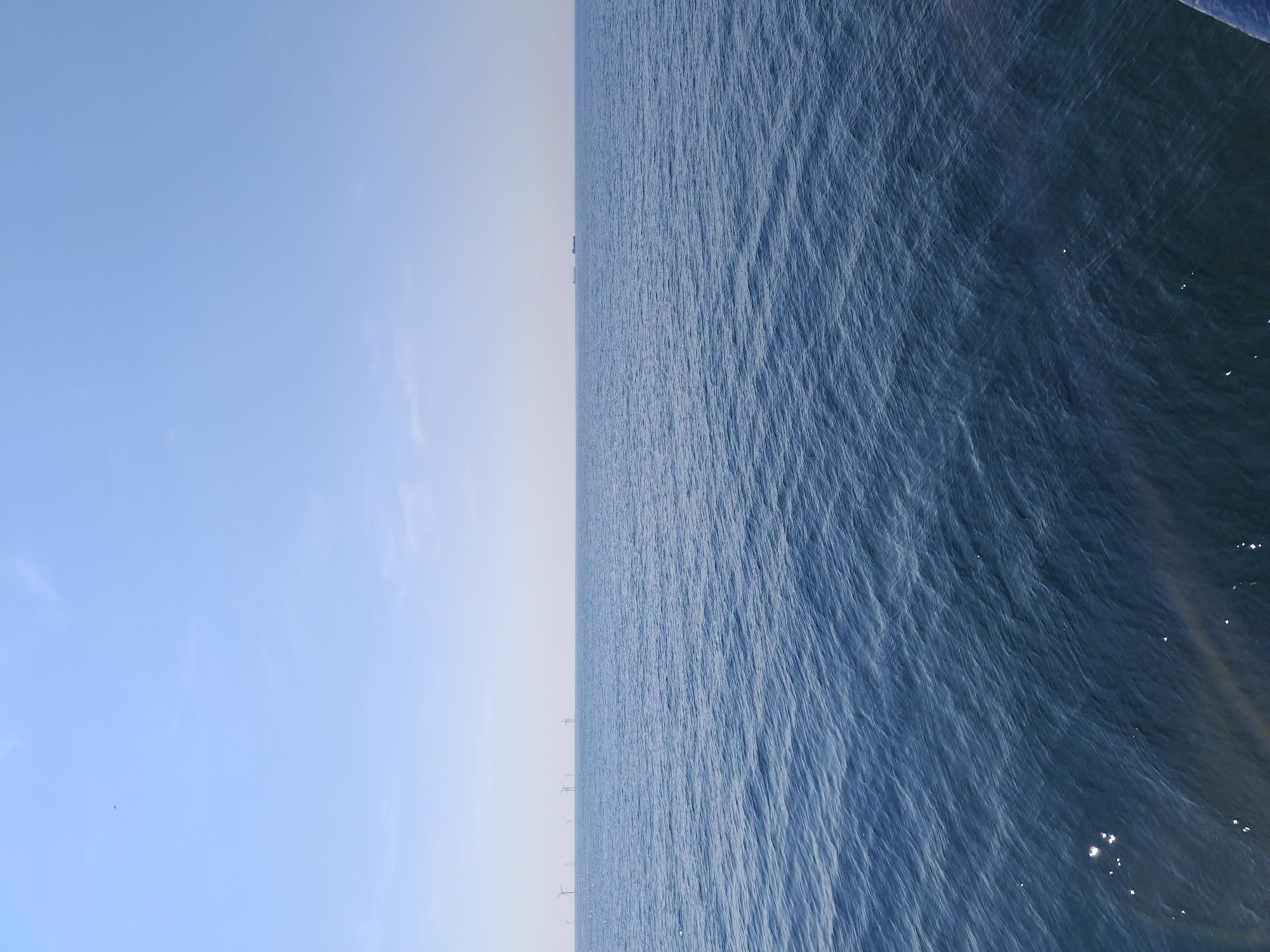 The image shows a view of the sea. On the left, a light blue sky is visible. Towards the center, the sky transitions into a hazy, light gray color, meeting the horizon. From the horizon stretching towards the right, the scene is dominated by the blue sea. The water shows gentle ripples and variations in tone, indicating movement and depth. In the distance along the horizon, several wind turbines are sparsely visible. Reflections of light are visible in the lower right corner.