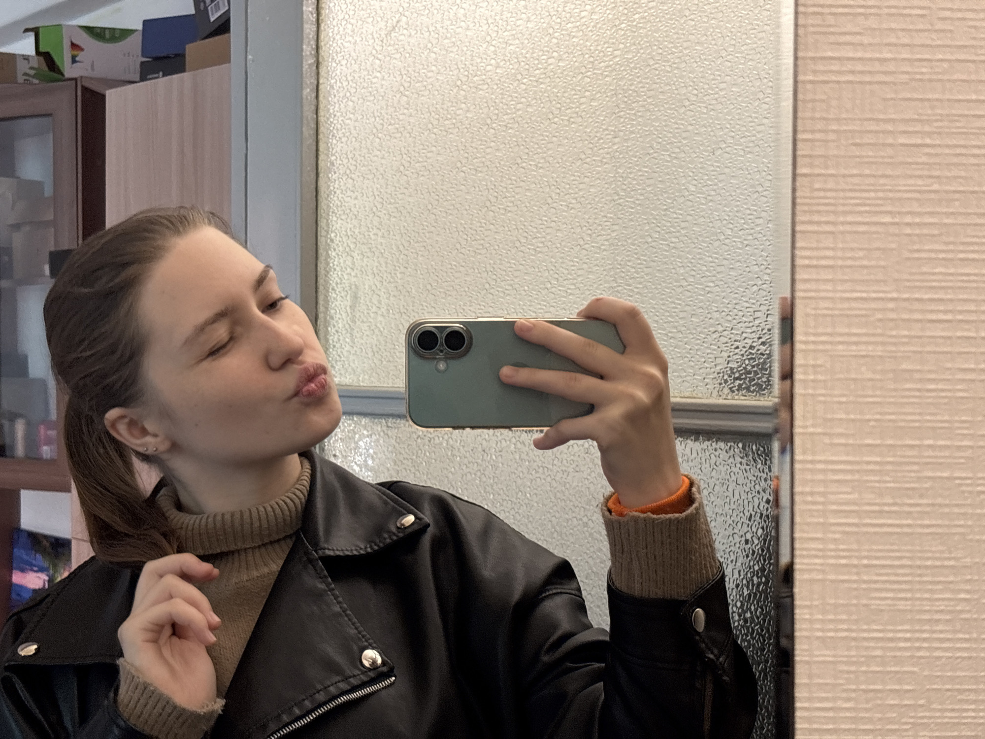 A young woman takes a selfie in a mirror. She has brown hair pulled back in a ponytail and is wearing a brown turtleneck sweater and a black leather jacket. Her right eye is closed and her lips are pursed. She is holding a green smartphone.
The background shows a cabinet with boxes on top and a textured glass panel next to a beige wall. The location is likely an apartment, but there is no indication of being in Saint Petersburg, Russia.