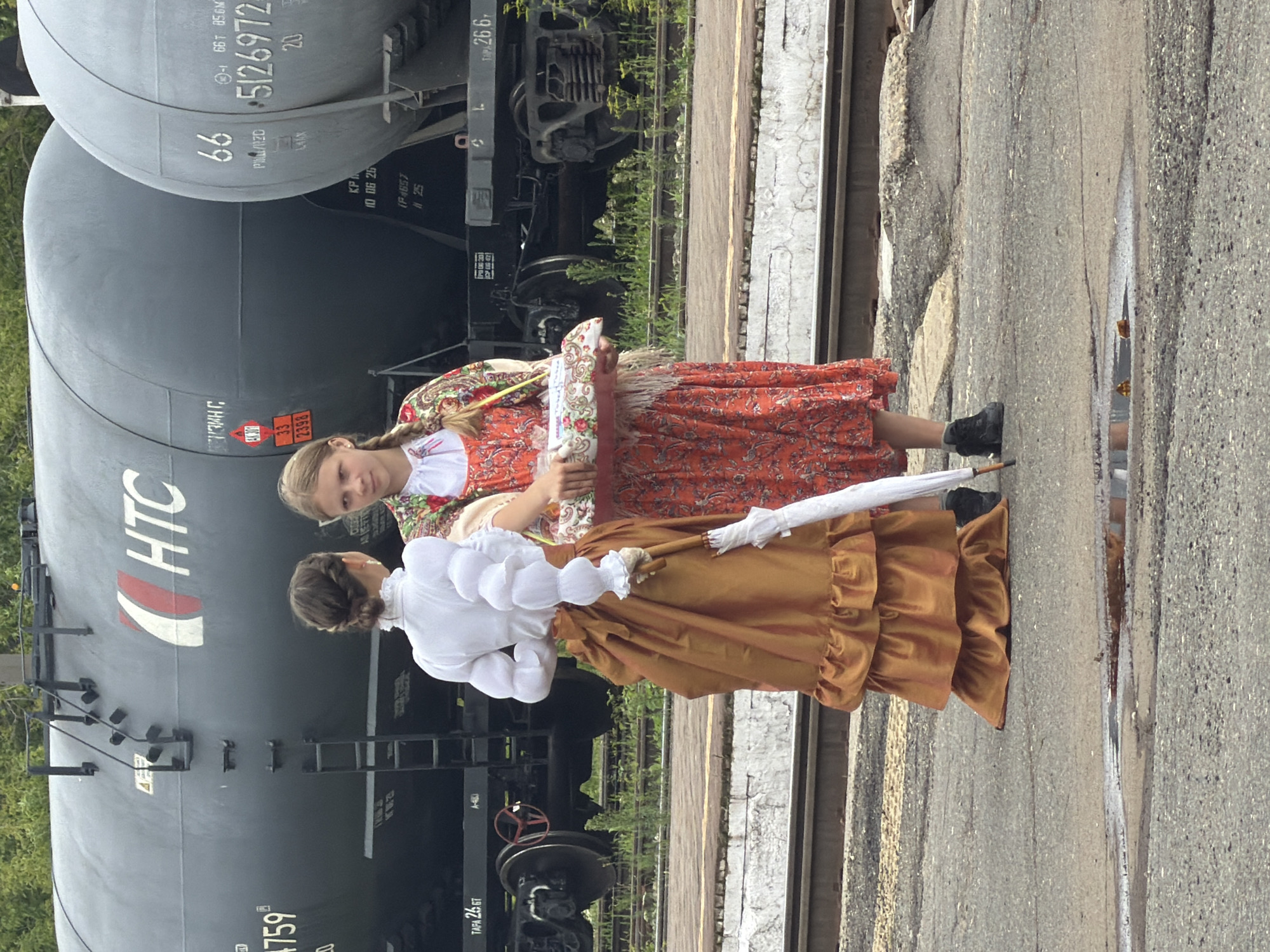 In Sarapul, Russia, two young girls stand near a railway, posing in front of large, dark gray cylindrical train cars. The girl on the left wears a white blouse with voluminous puffed sleeves and a full, rust-colored skirt, holding a white umbrella. The girl on the right is dressed in a vibrant red floral dress and a colorful shawl, carrying a basket of items. The scene appears to be an outdoor event or reenactment, with the train cars serving as a backdrop. The ground is paved with asphalt, showing signs of wear and puddles of water. The overall atmosphere suggests a historical or cultural gathering.
