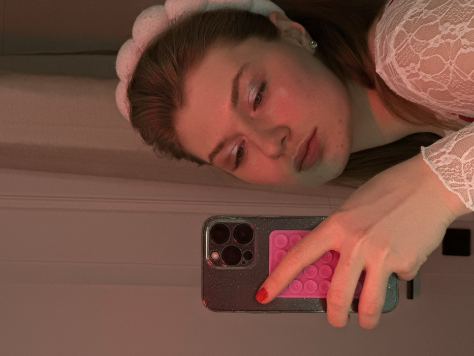 A woman poses in a selfie, wearing a lacy white top and a fluffy white headband. She looks downward with a slightly melancholic expression. Her hand holds a smartphone, which is reflecting the overhead lighting. The phone has a sparkling grey case and a pink, round-bubble grip on the back. The lighting creates a red-toned hue throughout the image.