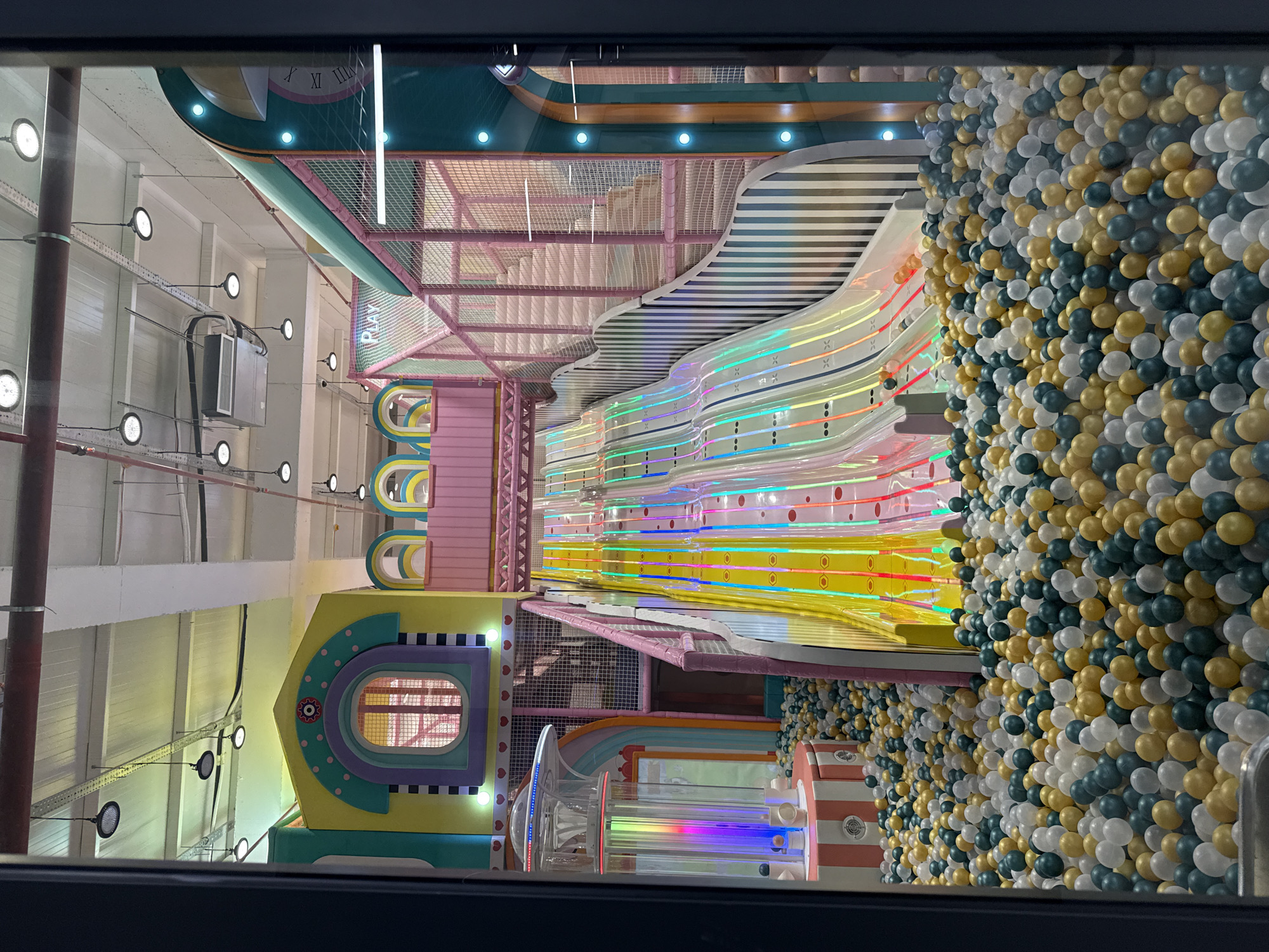 The image shows a children's indoor playground in Batumi, Georgia. The playground features a ball pit with white, gold, and dark teal balls. There are also slides with colorful LED lighting, and a play structure with pink, yellow, and teal elements, including a window and striped patterns. The ceiling above has industrial lighting fixtures. A decorative clock is also visible on a teal structure in the top center. The scene is viewed through a dark frame, suggesting it was taken from outside the playground's enclosed area.