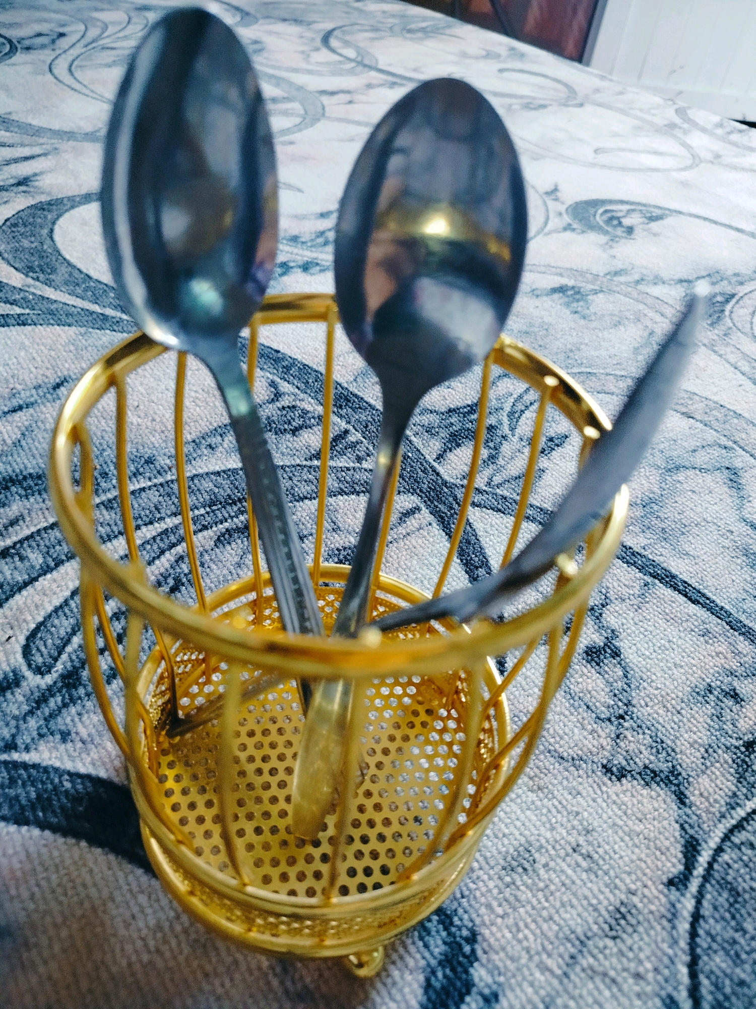 A close-up, eye-level shot captures a gold wire utensil holder containing three metal spoons. The holder is cylindrical with an openwork design, and it rests on a patterned surface. The background is blurred but appears to be a table or countertop with a fabric tablecloth exhibiting a grey and white swirling pattern. The lighting is moderate, creating some reflections on the spoons. The scene is indoors and suggests a domestic setting. There are no people visible in the image. It is not possible to determine the location in the city of Көлкент, Kazakhstan, from the visual information provided.