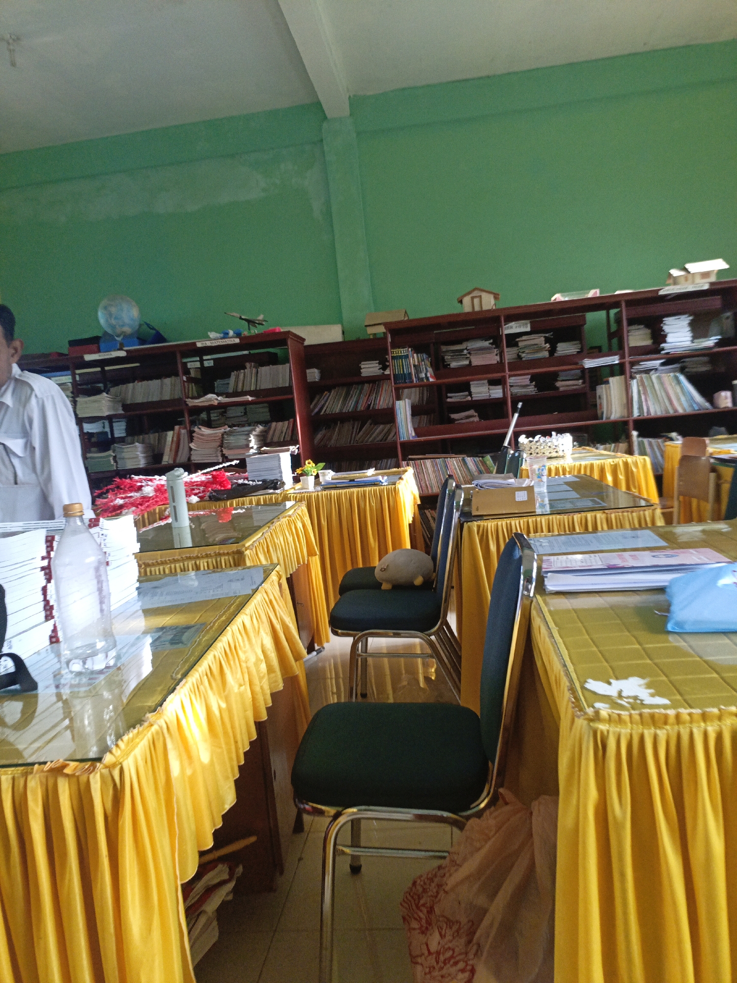 The image shows an indoor scene, likely a classroom or a library in Lhokseumawe, Indonesia, with green painted walls and wooden bookshelves filled with books and papers. Several tables are covered with bright yellow tablecloths, and some are topped with glass. There are chairs around the tables, with one chair in the foreground featuring a small, plush toy. The scene suggests an organized, though slightly cluttered, educational or administrative environment.