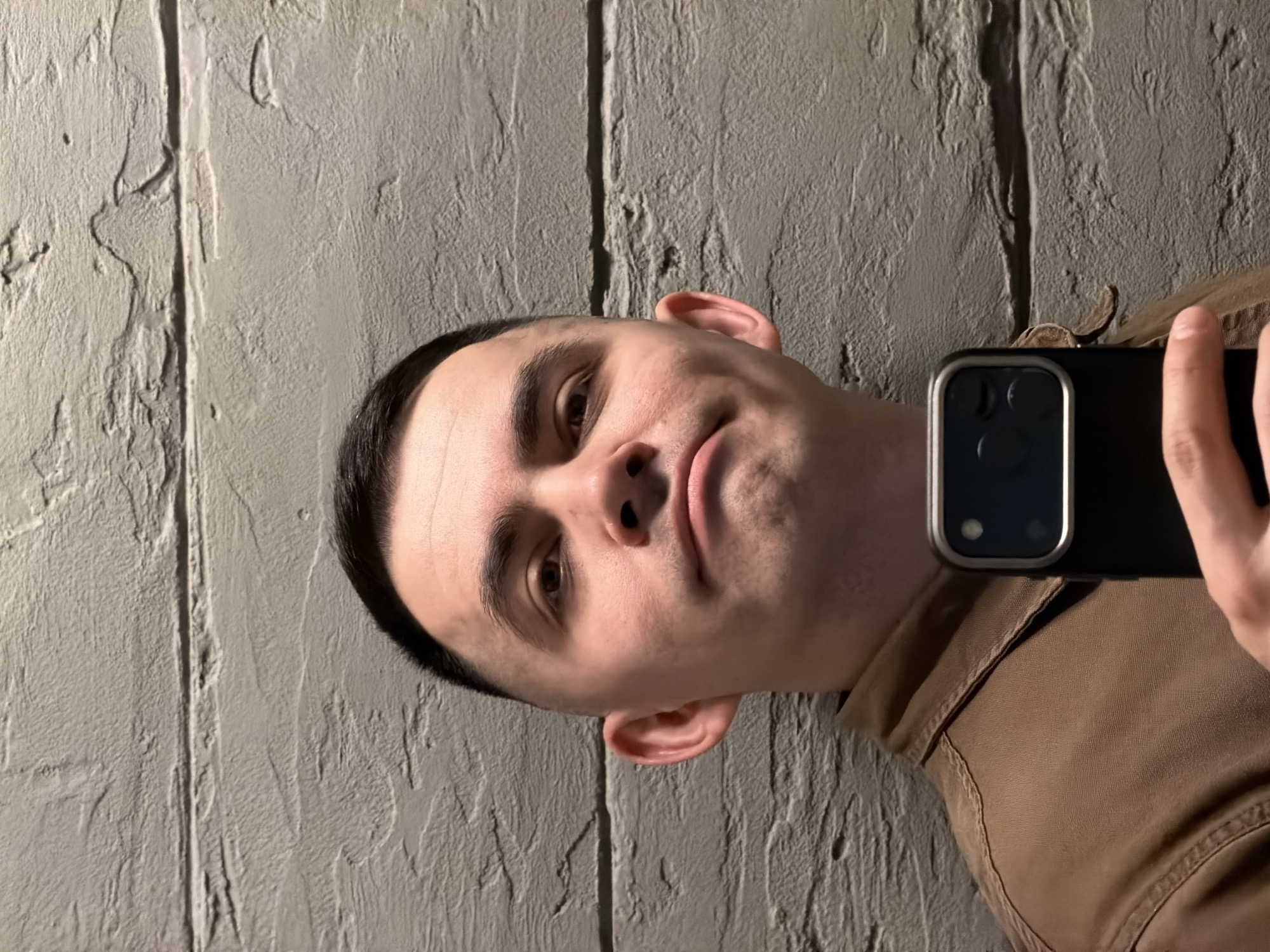 This portrait-style image, rotated 90 degrees clockwise, captures a young man taking a selfie indoors. The subject, a man with light skin, short dark hair styled in a buzz cut or fade, dark eyebrows, and dark eyes, looks directly at the camera. He has a subtle, almost smirking expression with his lips slightly parted.
He is holding a black smartphone, likely an iPhone Pro model due to the visible three-camera array and metallic frame, in his right hand. A portion of his right hand and thumb is visible as he grips the device. He wears a brown or tan-colored garment, possibly a jacket or shirt, with visible stitching and fabric texture, which is seen on the right side of the frame (due to the rotation).
The background consists of a textured wall, appearing to be light gray or beige-colored concrete or stucco, with a rough, uneven surface. A distinct vertical groove or seam runs through the wall behind him, creating a linear shadow and emphasizing the texture. The lighting is somewhat dramatic, casting shadows that highlight the wall's uneven surface and the contours of the man's face, suggesting an artificial light source indoors. There is no discernible text within the image. The scene is set in Izhevsk, Russia.