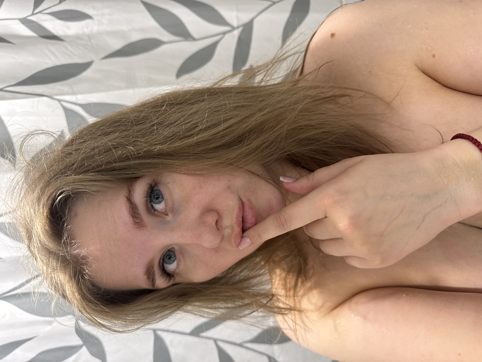 A young woman with light brown hair and blue eyes is lying down, possibly in a shower or bathroom setting, indicated by the shower curtain behind her. She is fair-skinned and has some water droplets on her skin. Her finger is touching her lips, and she is looking directly at the camera. A bracelet is visible on her wrist. The image does not provide sufficient information to confirm any details about the city of Ферма, Russia.