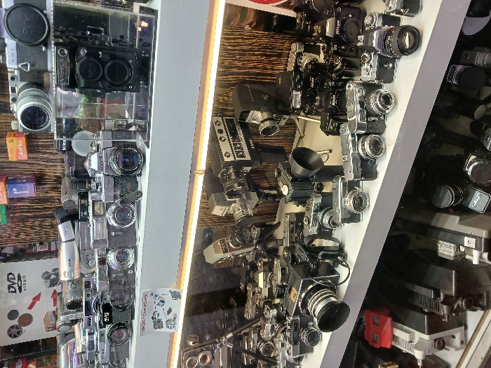 The image depicts a shopfront display in Istanbul, Türkiye, at the coordinates 41.022 latitude and 28.976 longitude. The display is located inside a shop, behind a glass window, and features a large collection of vintage cameras and other photographic equipment. The cameras are arranged on two shelves, one above the other, and are primarily silver and black in color. Some of the cameras are on stands, while others are stacked on top of each other. Several of the cameras have lenses pointed directly at the viewer, creating a sense of anticipation. The display also includes several other objects, such as a DVD video sign, and a variety of smaller items, including what appears to be an instruction manual for a camera. The overall impression is of a well-organized and carefully curated collection of vintage photography equipment. There are no people visible in the image.