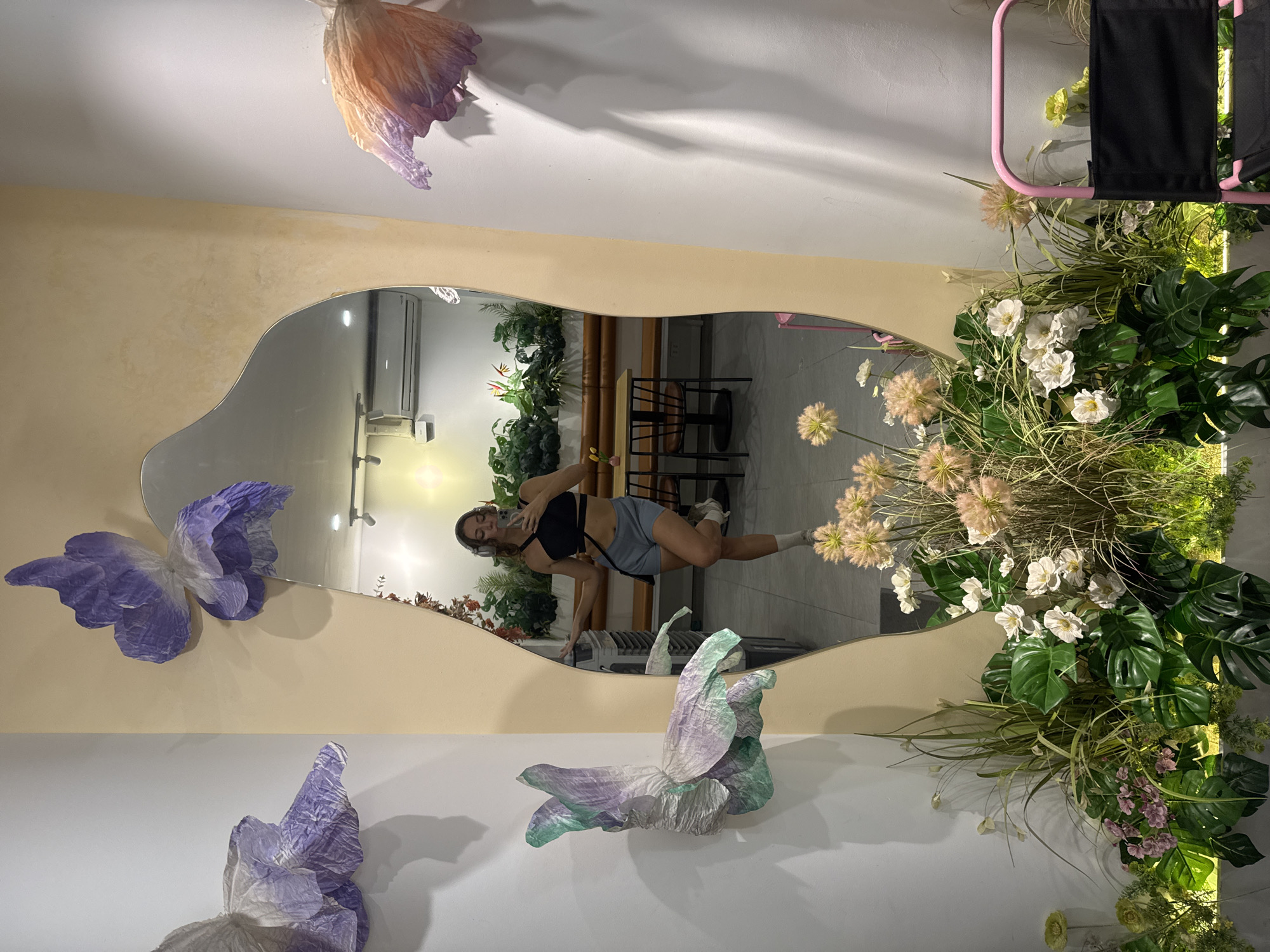 A full-body mirror reflects a young woman wearing headphones, a black crop top, and light blue shorts, holding her phone to take a selfie. She is standing in front of a wall decorated with large, abstract paper flowers in shades of purple, green, and orange. The mirror is framed by a beige, irregular shape, and a lush display of artificial plants and flowers, including large green monstera leaves and delicate white and pink blossoms, sits at the base of the mirror. The reflection in the mirror shows a restaurant or cafe interior with wooden tables and benches, and a wall covered in greenery. The overall setting appears to be a stylish, modern interior space, possibly a boutique or a cafe, with decorative elements that create a whimsical and artistic atmosphere.