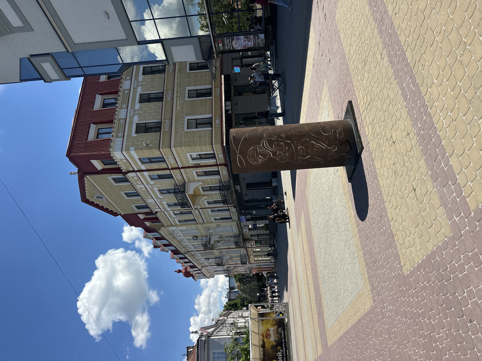The image shows a street scene in Ternopil, Ukraine, likely on a sunny day. In the foreground, a patterned pavement of red and yellow bricks leads towards a cylindrical sculpture with etched figures. Beyond the sculpture, a street lined with multi-story buildings unfolds. The buildings feature a mix of architectural styles, with some exhibiting classic European design elements like ornate facades, balconies, and red-tiled roofs, while others present modern glass exteriors. Several people are visible walking along the street, some browsing at market stalls, and others sitting on chairs. There are also awnings and storefronts indicating commercial activity. The sky is blue with scattered white clouds, and some electrical wires are visible against the sky. The overall impression is of a lively urban environment.