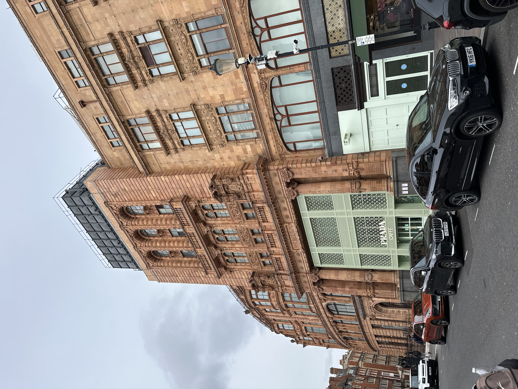 The image depicts a street scene in London, United Kingdom. The focal point is the ornate facade of a building, notable for its architectural details and featuring the "PRADA Caffe" storefront. Several parked cars line the street in front of the building, including black and orange vehicles. Pedestrians are partially visible further down the street. The sky is overcast, suggesting a cloudy day.