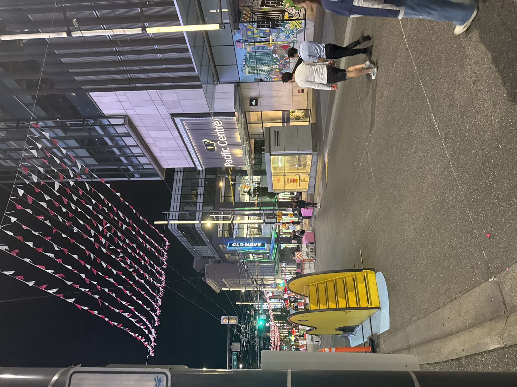 A nighttime street scene in Vancouver, Canada, captures a vibrant urban setting decorated with strings of red and white triangular flags overhead. The image focuses on the Pacific Centre, a large building with modern architecture, featuring glass facades and vertical metallic elements. A prominent sign reads "Pacific Centre" in illuminated lettering.
Adjacent to the Pacific Centre, a colorful mural depicting stylized city buildings and people adorns the exterior. In the foreground, a bright yellow, abstract structure with stairs is positioned on the street, possibly a temporary art installation or play structure. Several individuals are walking on the sidewalk and across the street. Two men in casual attire are walking away from the viewer towards the Pacific Centre. Further down the street, signs for "OLD NAVY" and "THOMPSON ST" are visible, indicating a commercial area. Traffic lights and streetlights illuminate the scene, casting a glow on the wet pavement. Orange traffic cones and pink barriers are also present, suggesting ongoing construction or street management. The overall atmosphere is lively and urban, despite the late hour.
