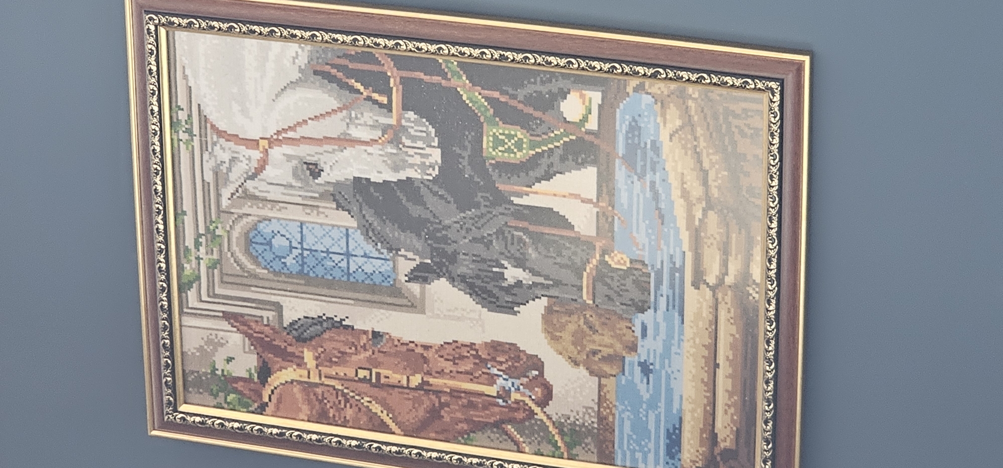 The image displays a framed cross-stitch tapestry, mounted on a blue wall. The photograph is taken at an angle, and the tapestry itself is rotated 90 degrees counter-clockwise.
When viewed in its intended upright orientation, the tapestry depicts a dynamic, classical-style scene. On the left, a majestic white horse is rearing on its hind legs, with a figure in dark attire visible on its back. In the lower right foreground, a brown horse is positioned, facing towards the left. Both horses are adorned with harnesses.
In the background, behind the horses, stands an ornate building featuring columns or arches and a prominent rounded window with blue-toned, possibly stained-glass, details. Green vines are shown climbing the building's exterior. Adjacent to the building, on the right side of the tapestry, a waterfall or significant water feature flows downwards.
The cross-stitch medium gives the entire artwork a distinct pixelated texture. The tapestry is enclosed within an elaborate frame, which is dark brown with intricate gold decorative elements along its inner edge. No visible text is present in the artwork or on the frame. The item is located in Manilva, Spain.